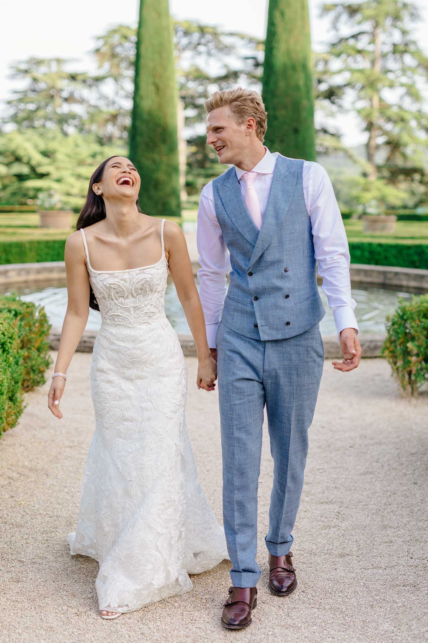 Bride and groom laughing while walking hand-in-hand along a garden path lined with cypress trees and hedges