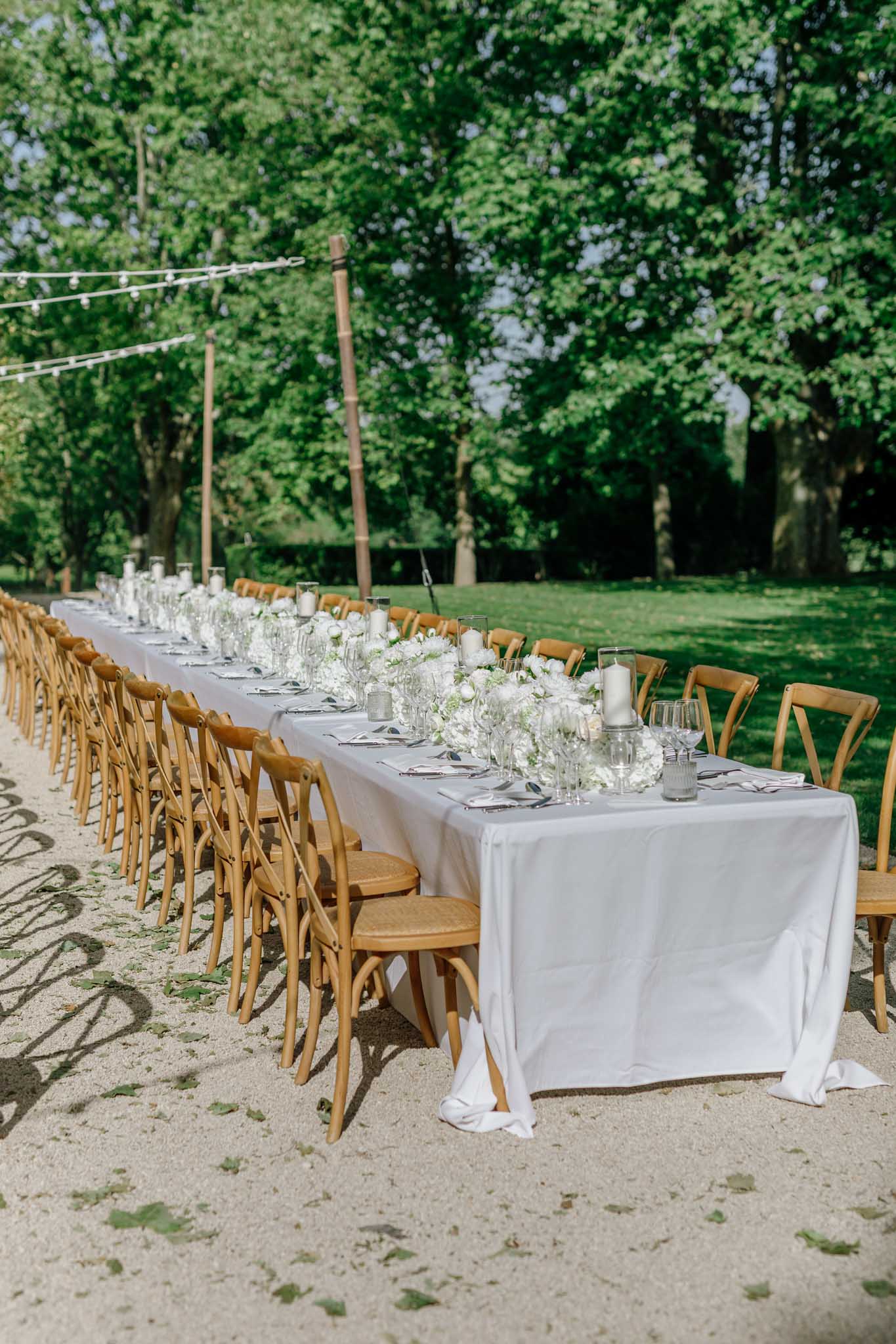 Outdoor reception table with white linen, glass vases of white flowers, and cross-back chairs under string lights and trees