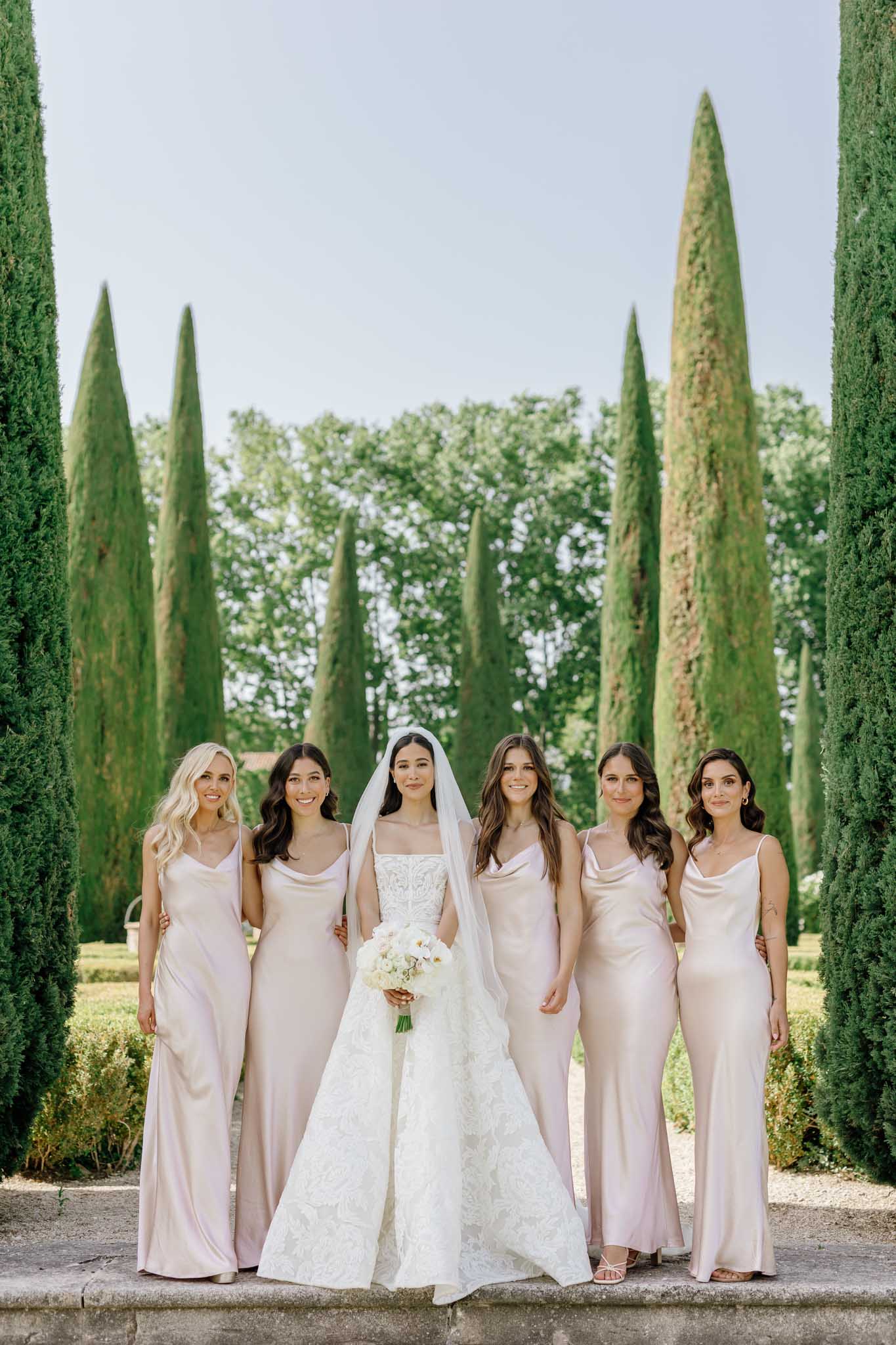 Bride with five bridesmaids in champagne silk dresses posing on garden path lined with Italian cypress trees