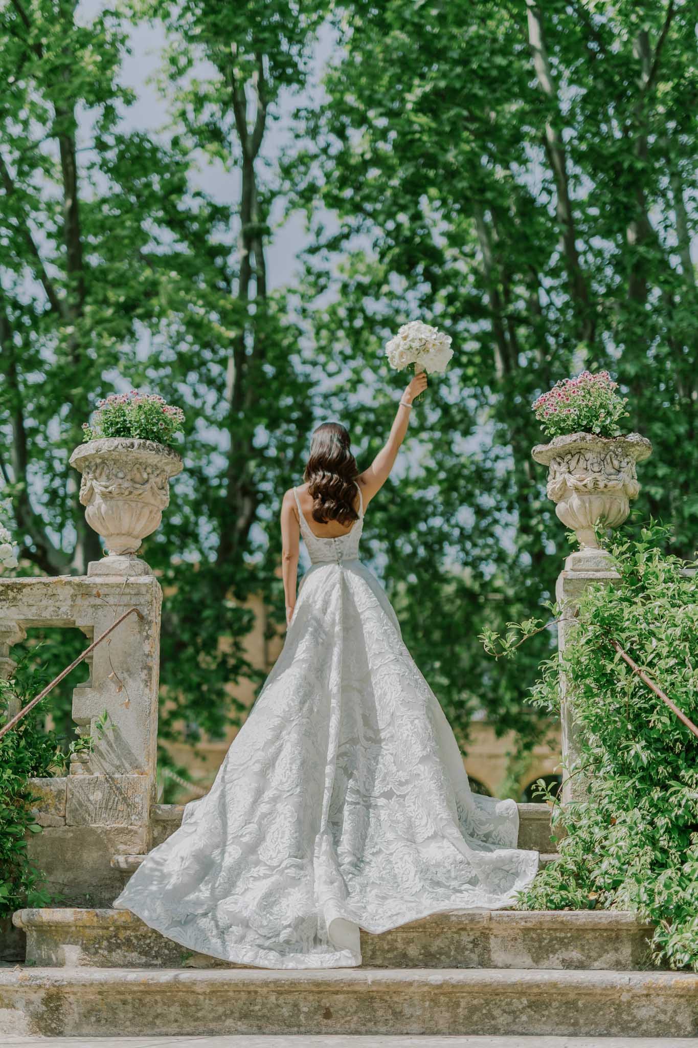 Bride raising bouquet on stone terrace flanked by stone urns with pink baby's breath at Chateau de Sannes