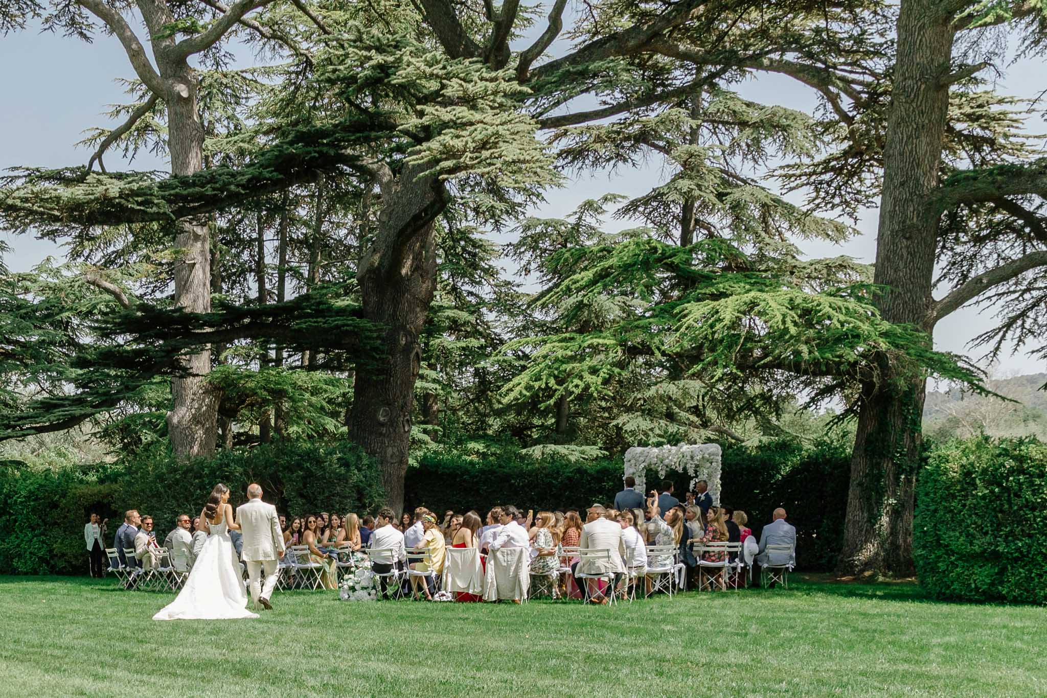 Outdoor garden ceremony beneath mature trees with bride and groom at a white-framed altar and seated guests