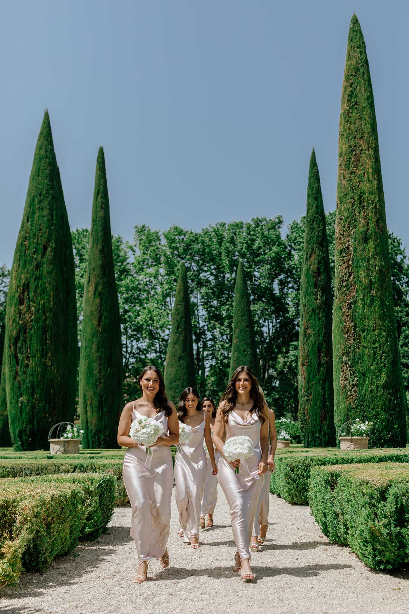 Bride and three bridesmaids in ivory slip dresses on cypress-lined garden path carrying white peony bouquets