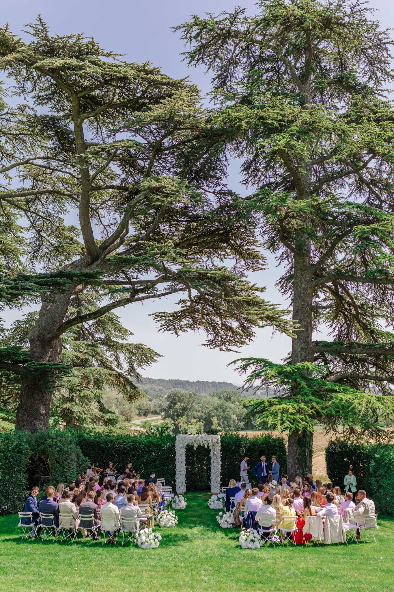 Outdoor wedding ceremony beneath cedar trees with 80-100 guests, white floral arch, and rolling countryside backdrop