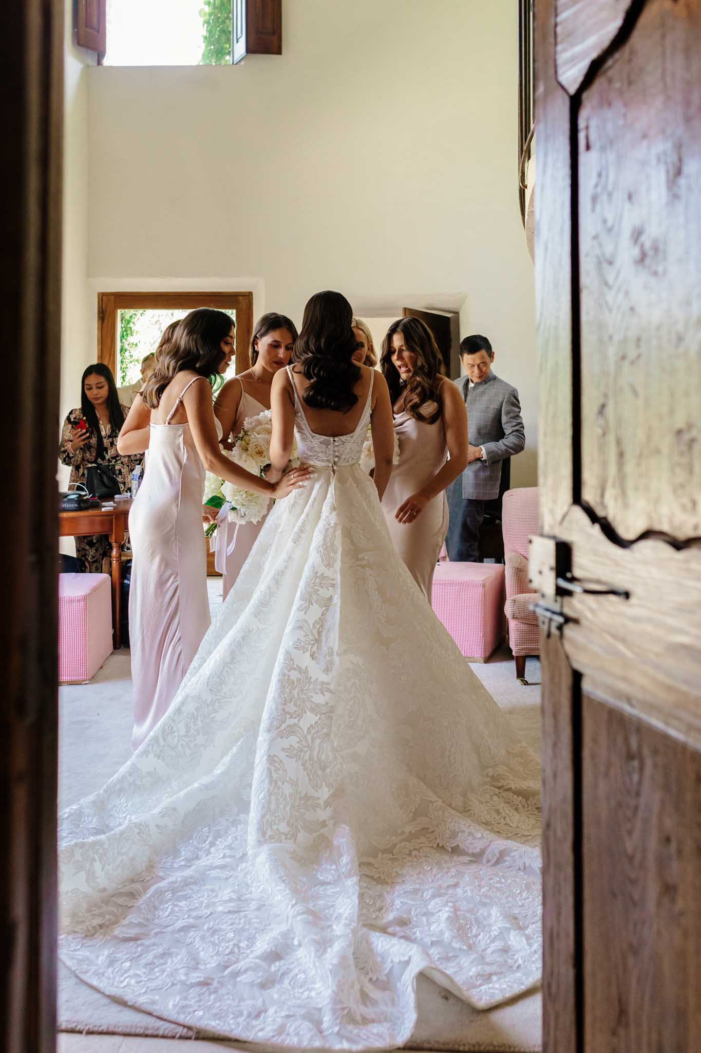Bride in ivory beaded A-line gown surrounded by bridesmaids in pale pink in rustic preparation room with pink poufs