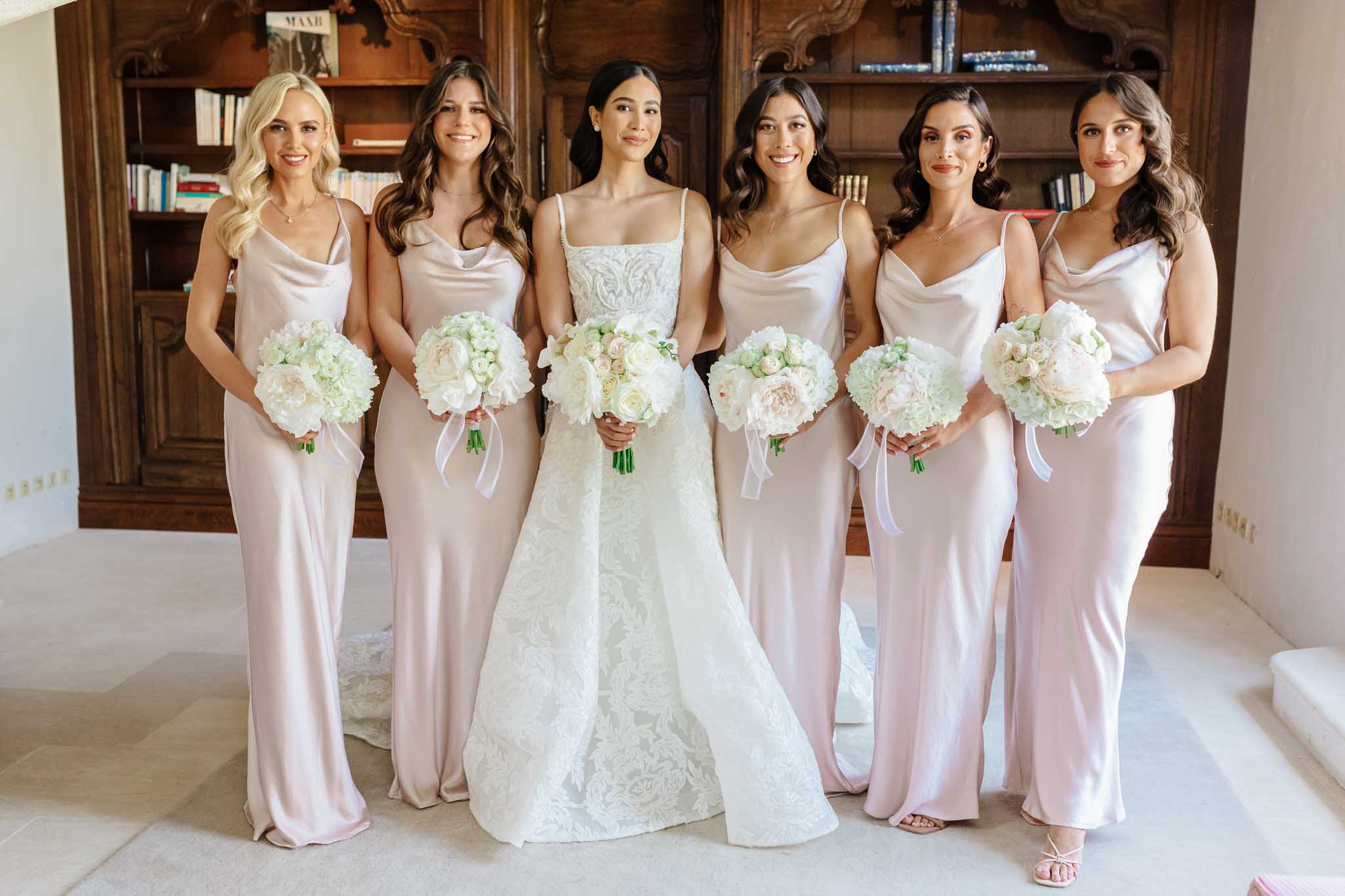 Bride in ivory lace gown flanked by five bridesmaids in blush pink satin dresses