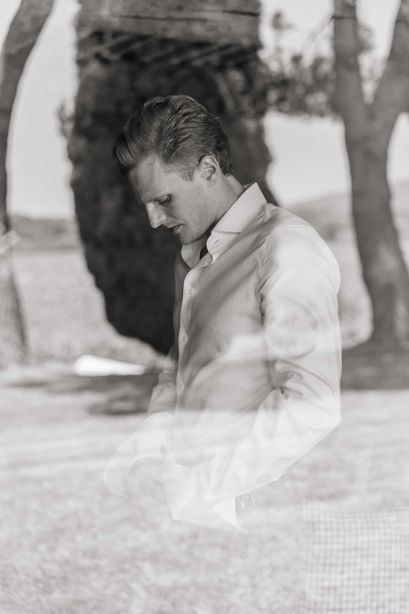 Groom in white dress shirt looking down in quiet contemplation during the ceremony, stone archway visible behind.