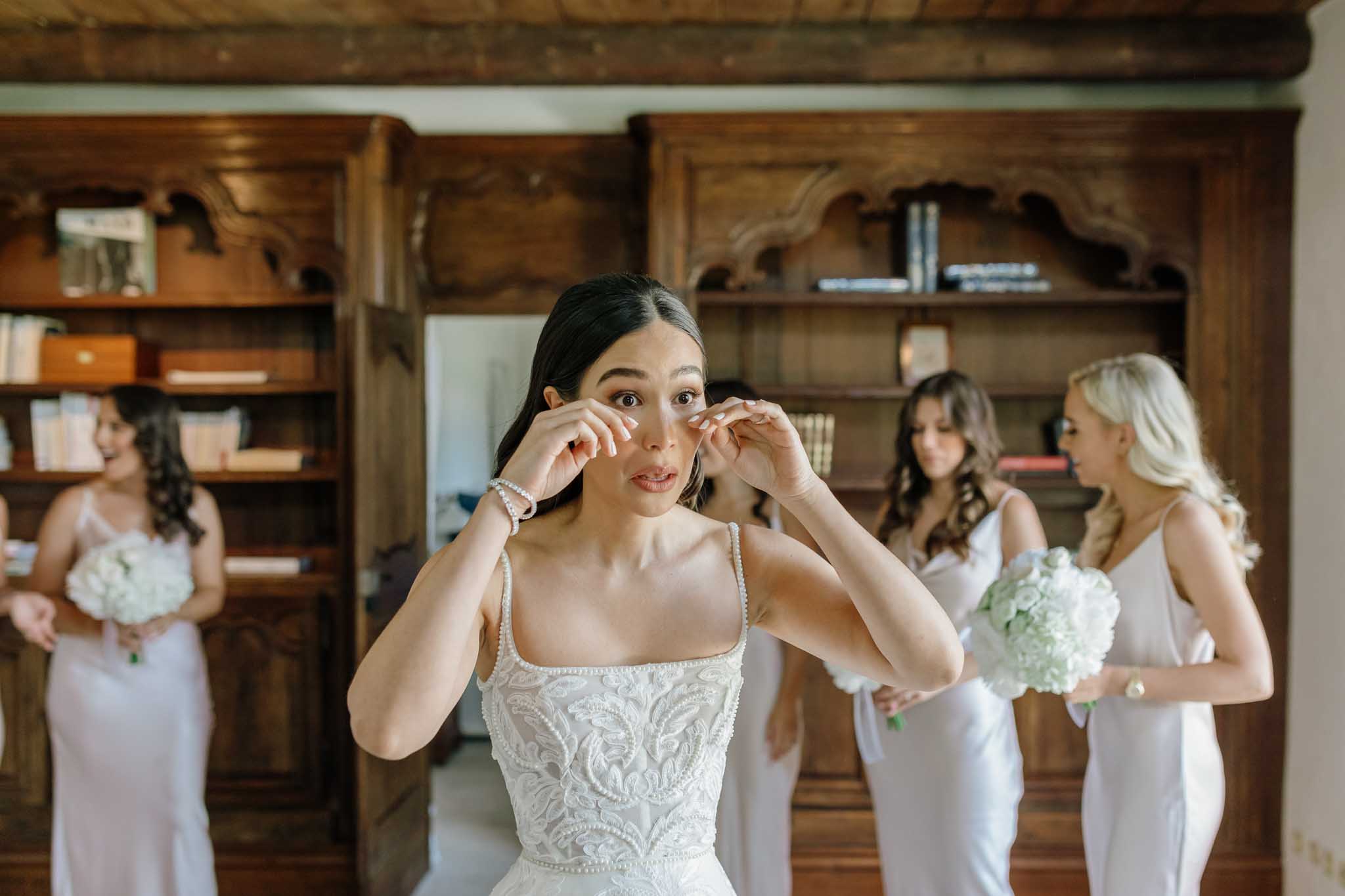 Bride adjusting earrings in lace wedding dress with three bridesmaids in lavender gowns in library setting