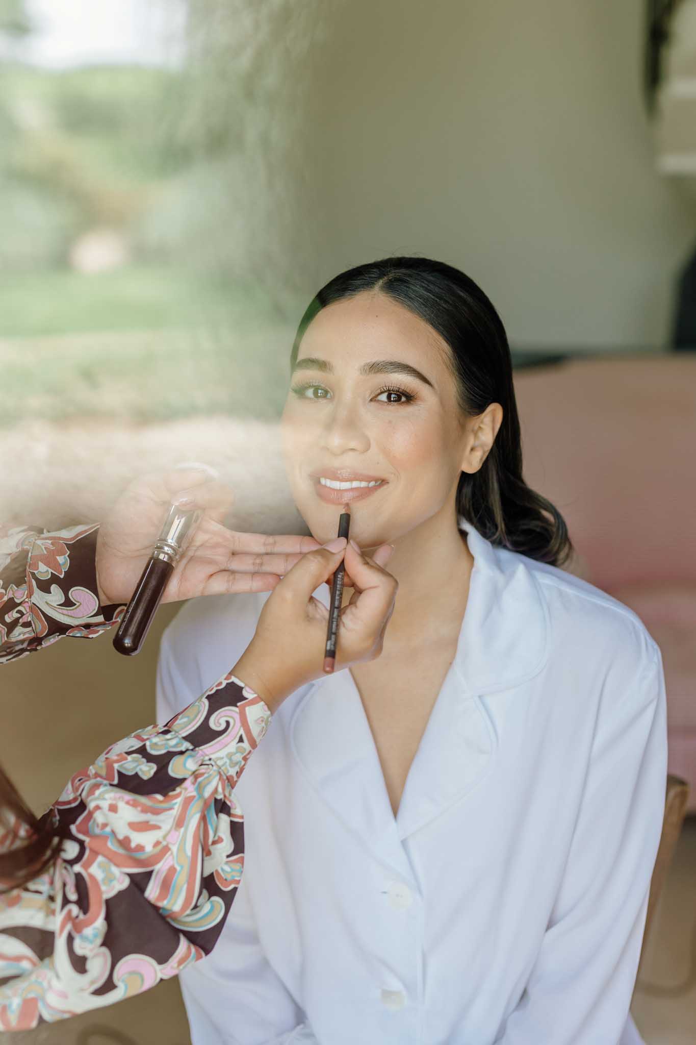 Bride in white robe smiling as makeup artist applies lip color during wedding morning preparations in neutral indoor space