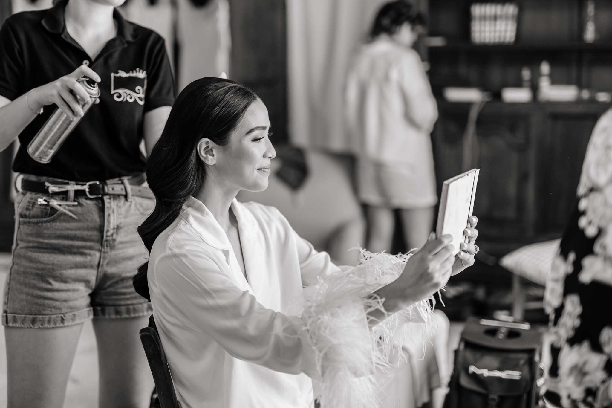 Black-and-white portrait of bride in white shirt holding handheld mirror while hairstylist applies hairspray