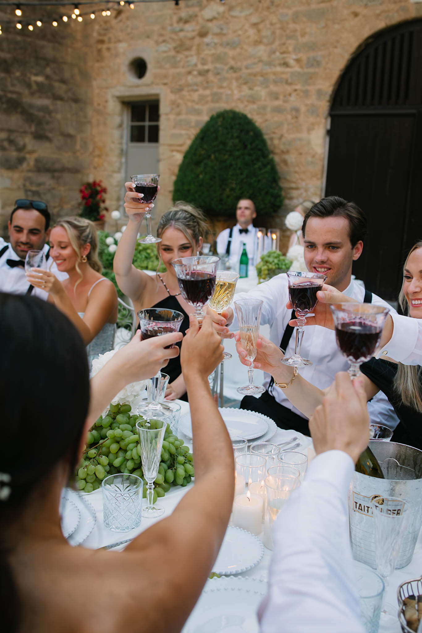 Wedding guests raise glasses for toast at long white-draped table in stone courtyard with string lights