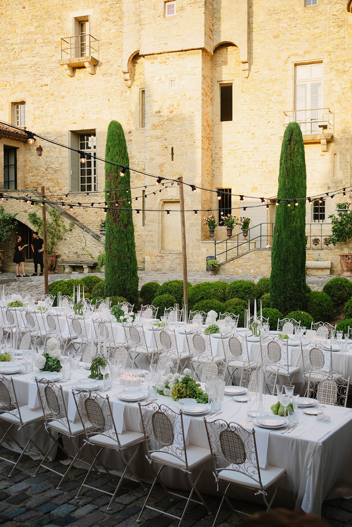 Outdoor reception in historic stone courtyard with long white-clothed tables, wrought-iron chairs, cypress topiaries, and black string lights