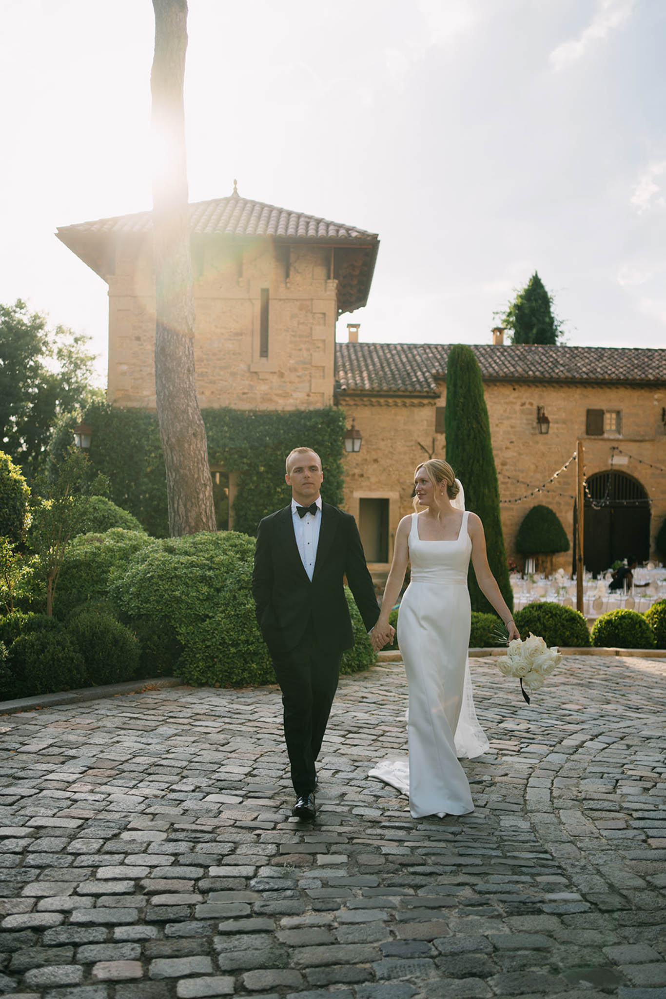Bride and groom walk hand-in-hand across stone courtyard in front of Tuscan-style villa with tower and cypress trees