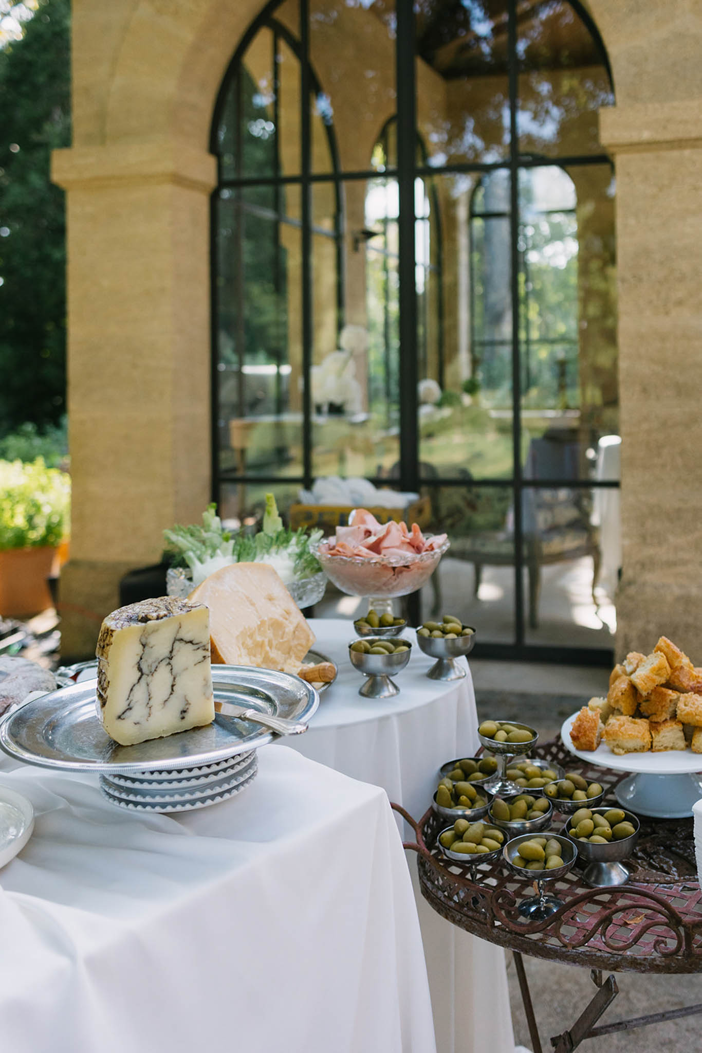 Cocktail hour cheese and charcuterie spread on white linen tables in front of stone archway with garden courtyard