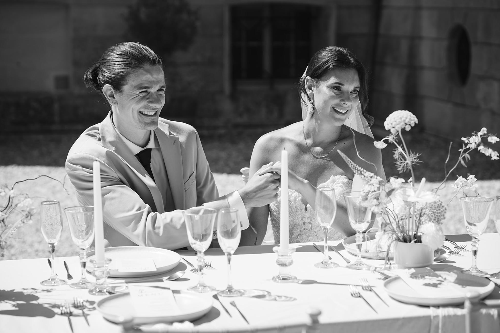 Black and white photo of bride and groom laughing at reception table with white taper candles and floral arrangements