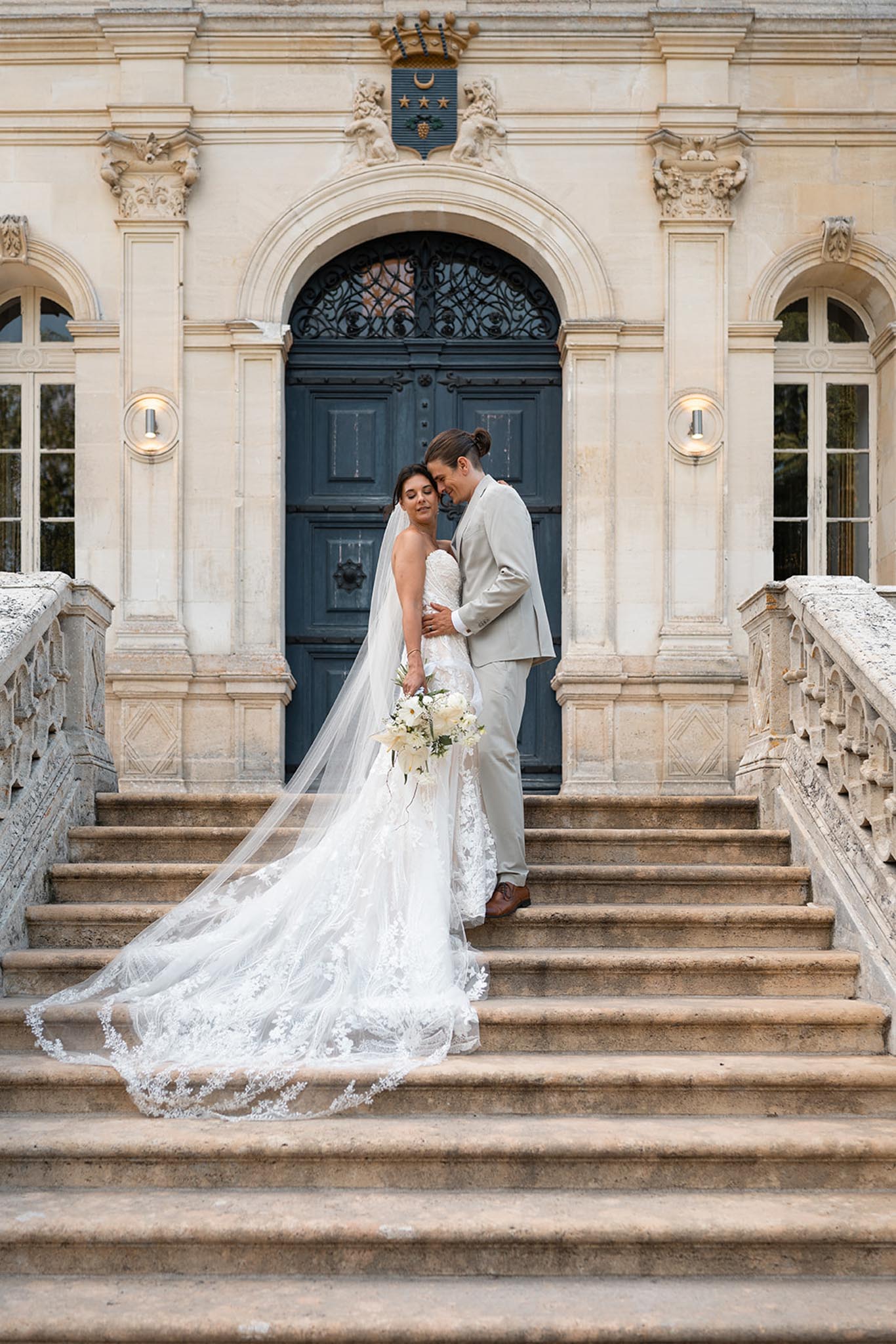 Bride and groom embrace on stone staircase before neoclassical faÃ§ade with columns and arched blue doorway