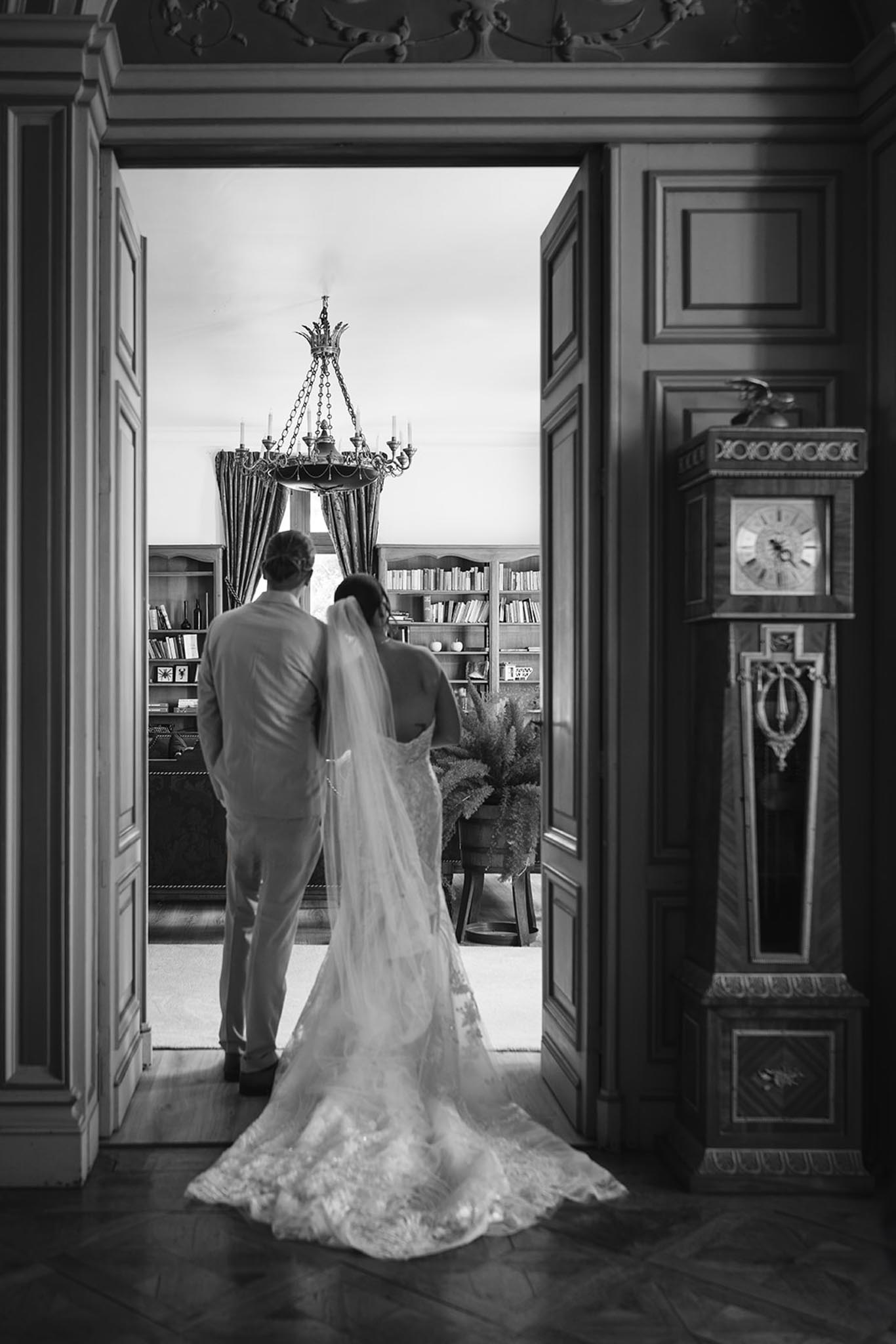 Black-and-white portrait of bride and groom from behind gazing into ornate interior with chandelier and bookshelves