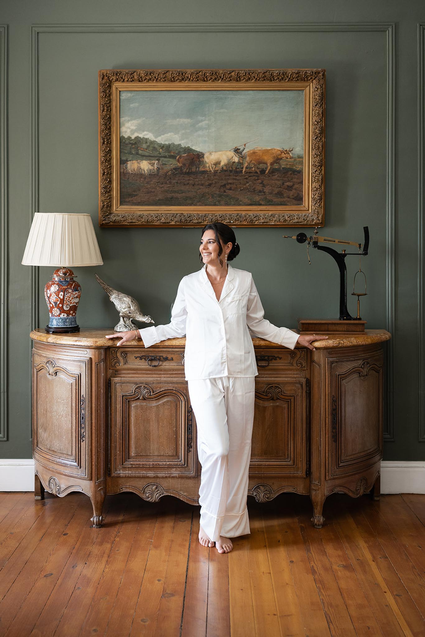 Bride in ivory linen pantsuit beside antique console table in classically furnished room with gilt-framed painting