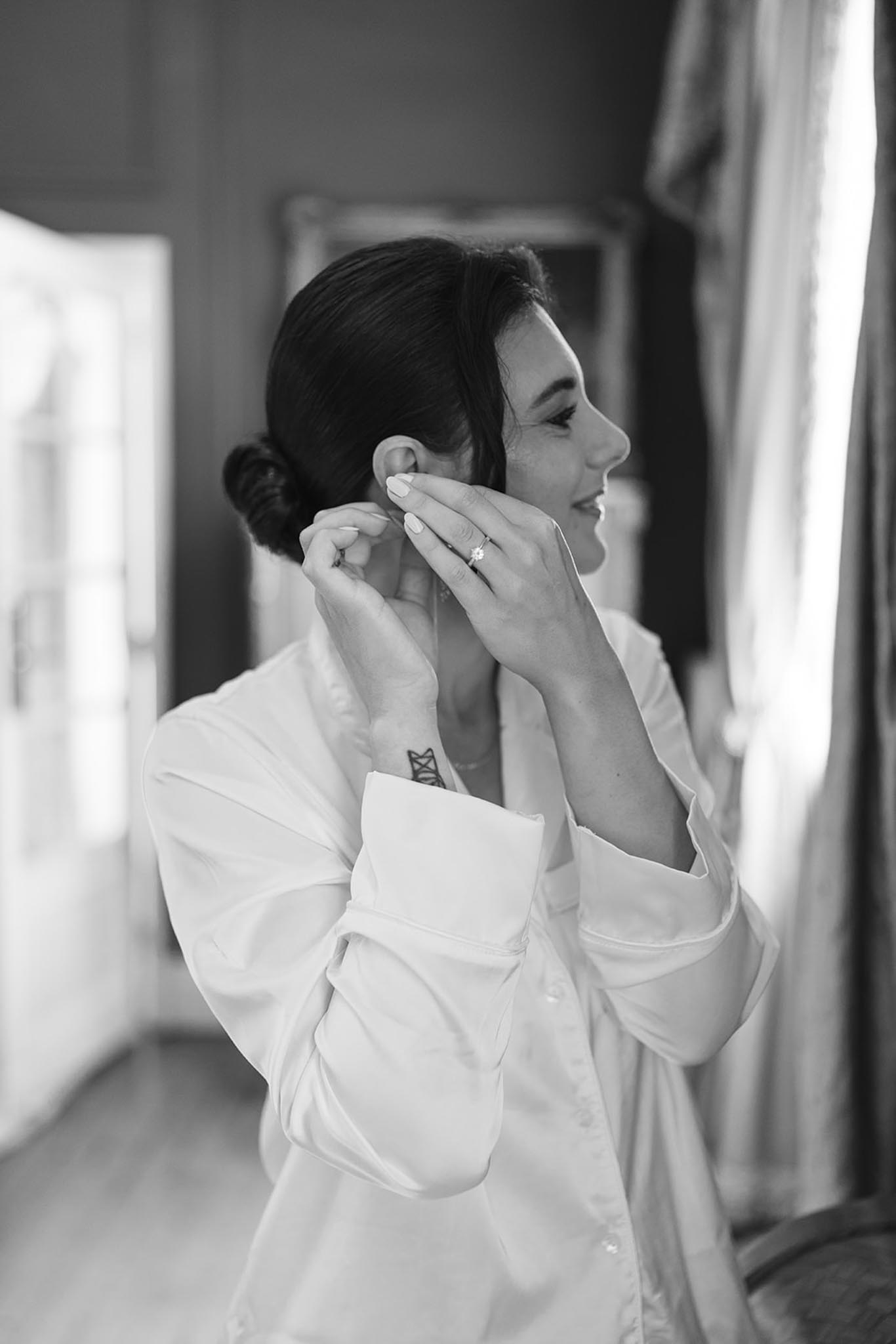 Black and white portrait of bride in white robe fastening earring, low updo, natural light from window, hallway behind