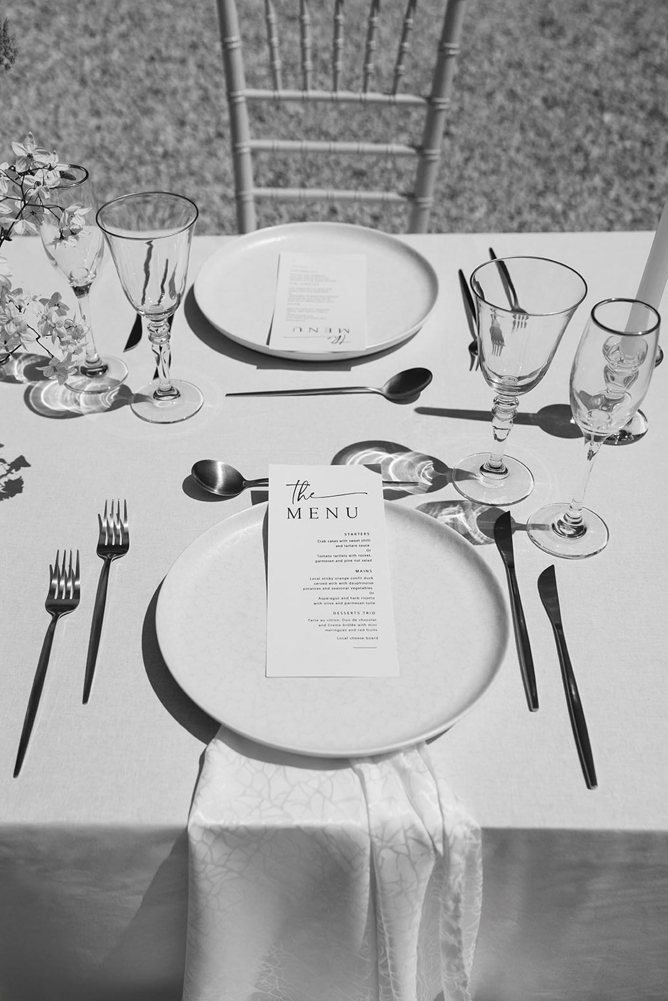 Black and white overhead shot of a formal place setting with layered plates, black flatware, and printed menu card