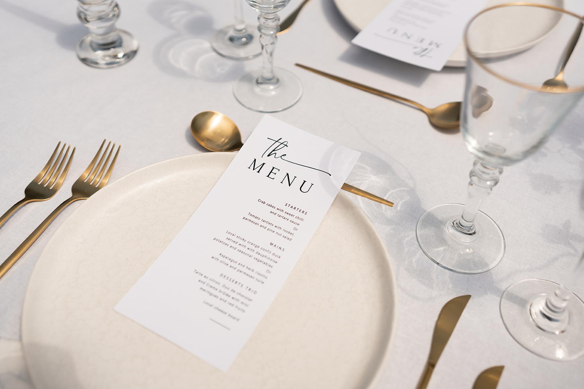 Formal table setting with white menu card on cream plate, gold flatware, and crystal glasses on white tablecloth