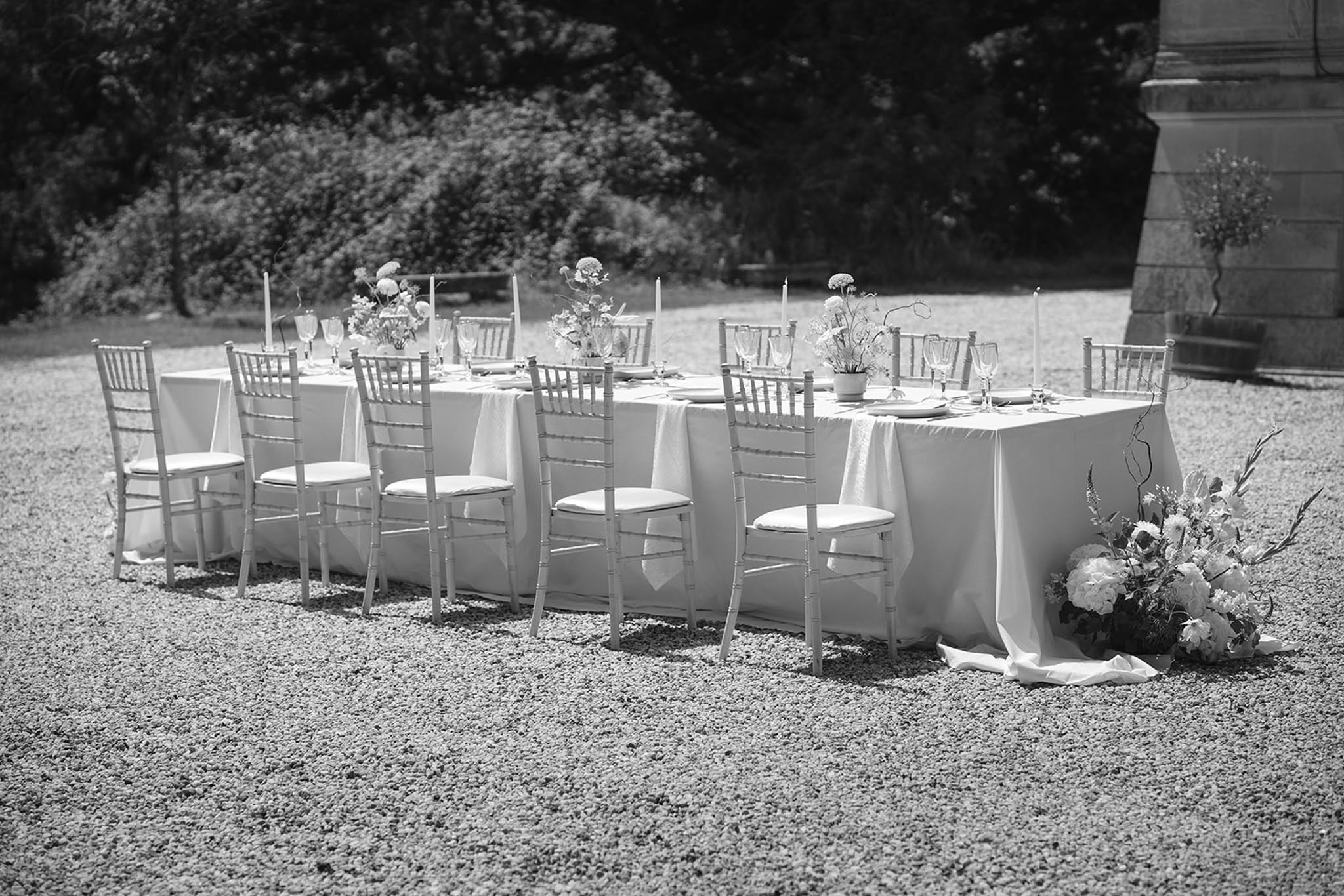 Black-and-white wide shot of two long outdoor tables with chiavari chairs and floral centerpieces in stone courtyard