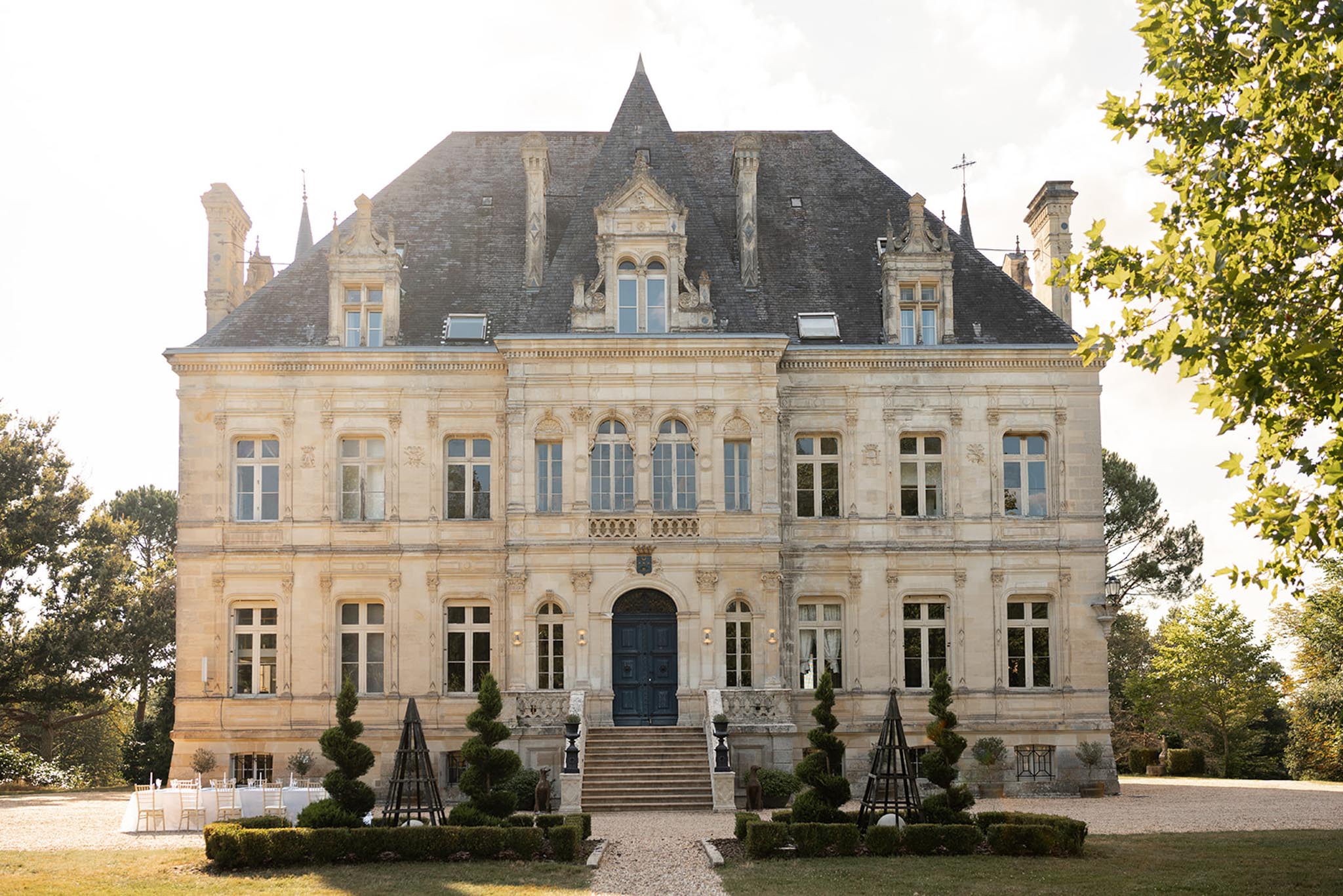 19th-century limestone chÃ¢teau with mansard roof and white Chiavari chairs arranged on gravel courtyard for ceremony