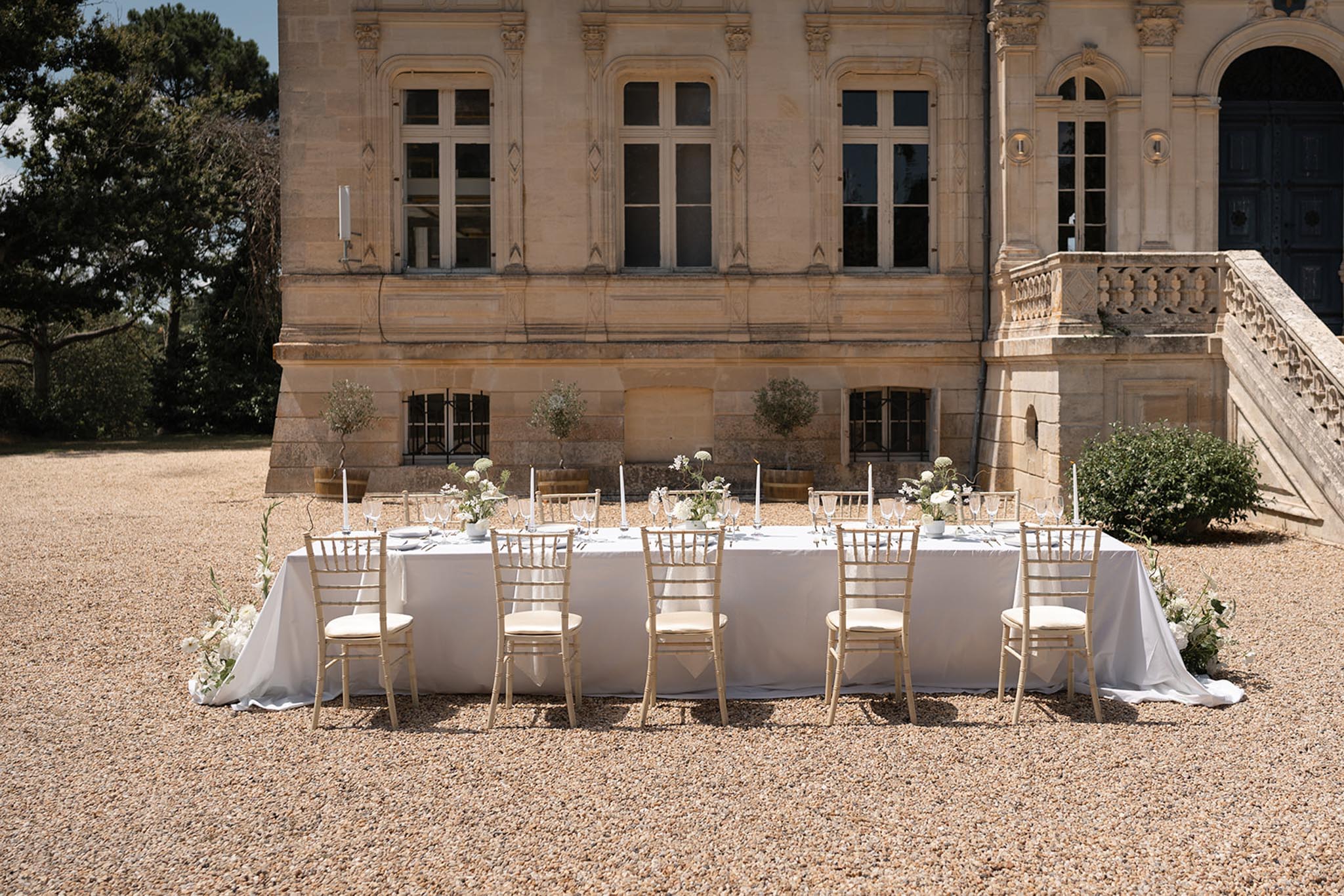 Single long banquet table on gravel courtyard in front of honey-coloured chÃ¢teau with white hydrangea centerpieces