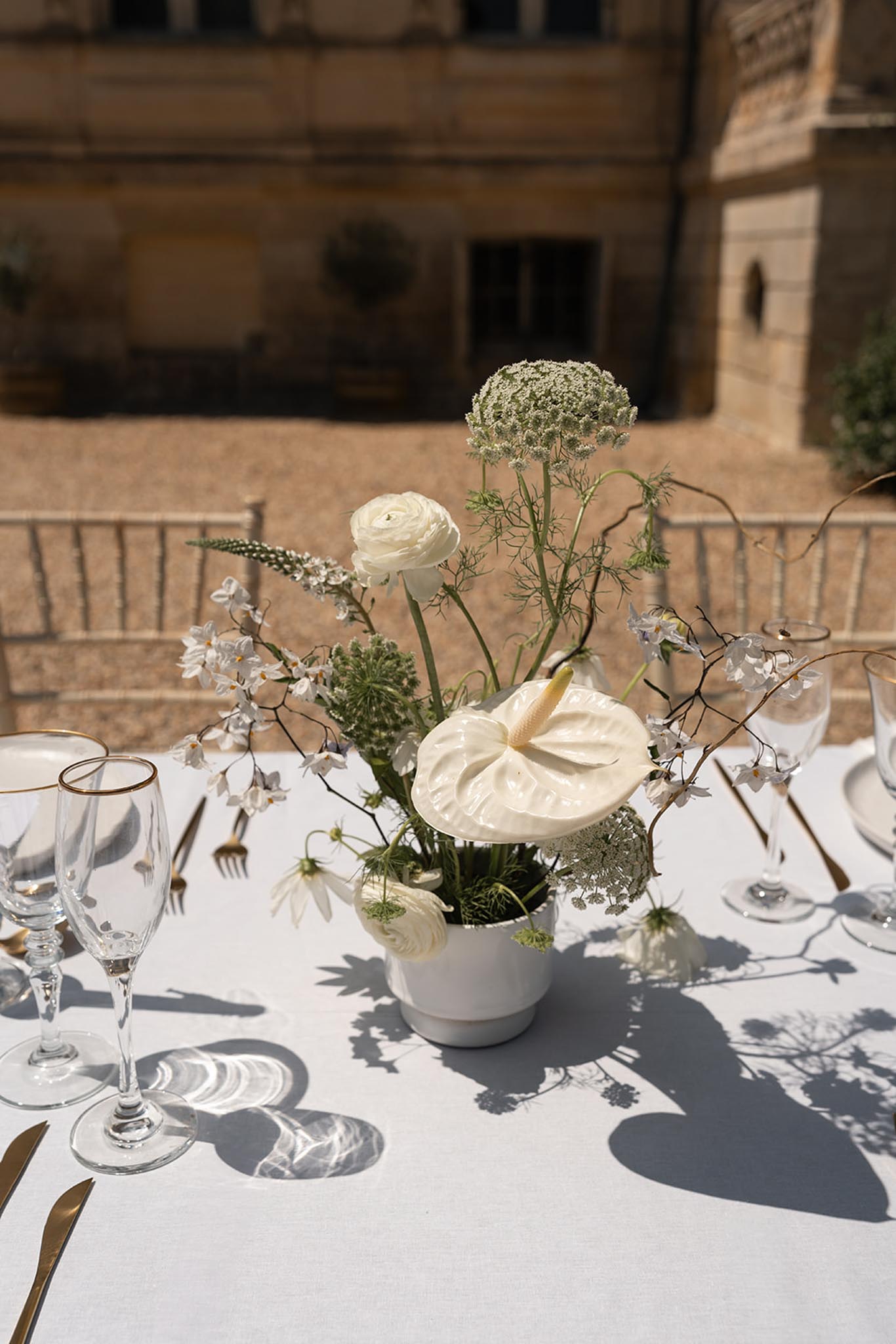 Reception centerpiece of white anthurium and ivory ranunculus in ceramic vase with gold-rimmed glassware beside it