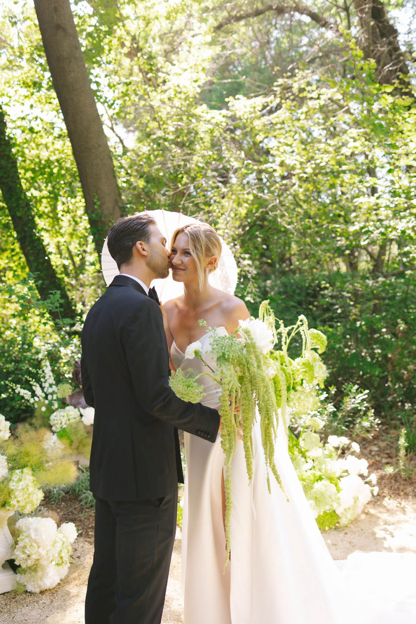 Groom in black tuxedo kissing bride on the cheek; bride in ivory gown holding a white hydrangea bouquet under a canopy of flowering trees.