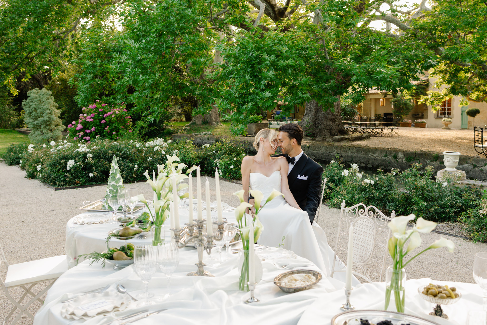 Bride and groom seated at a long white-linened table in a garden courtyard with candelabras and white floral centerpieces.