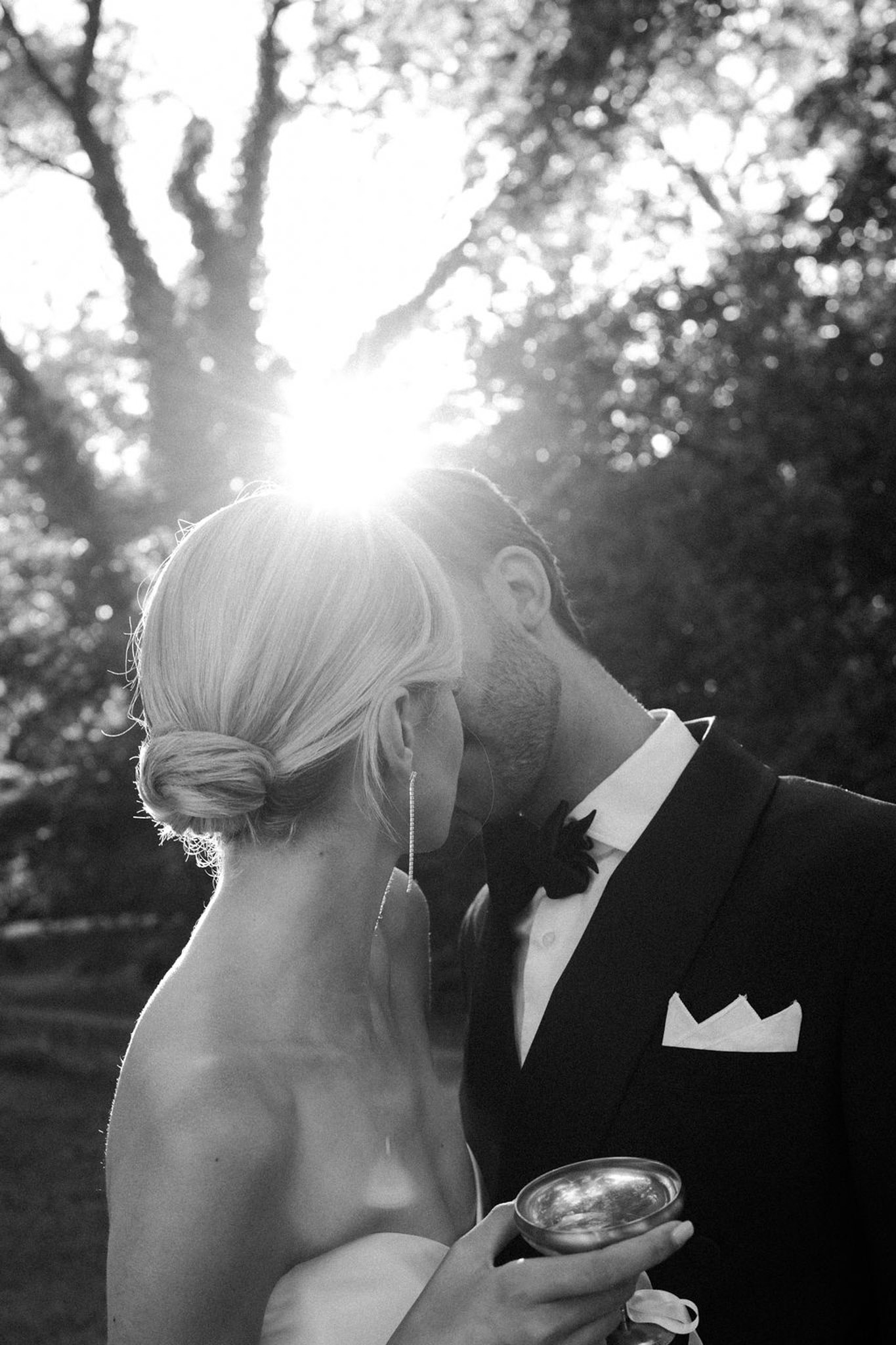 Black and white portrait of bride and groom in profile leaning close together, backlit by sunlight filtering through flowering trees.