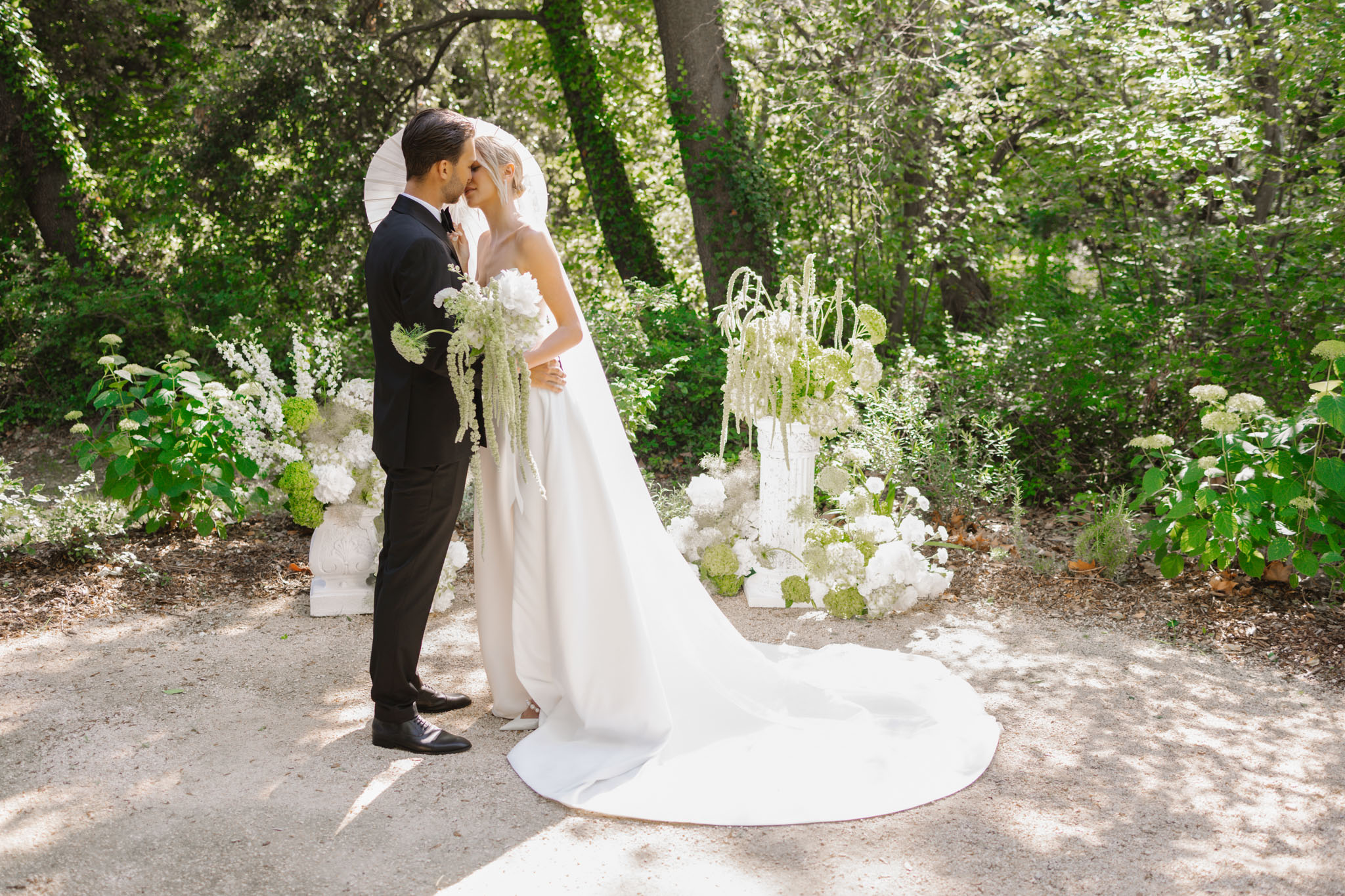 Bride and groom under a white umbrella on a garden path flanked by white hydrangea and amaranthus arrangements