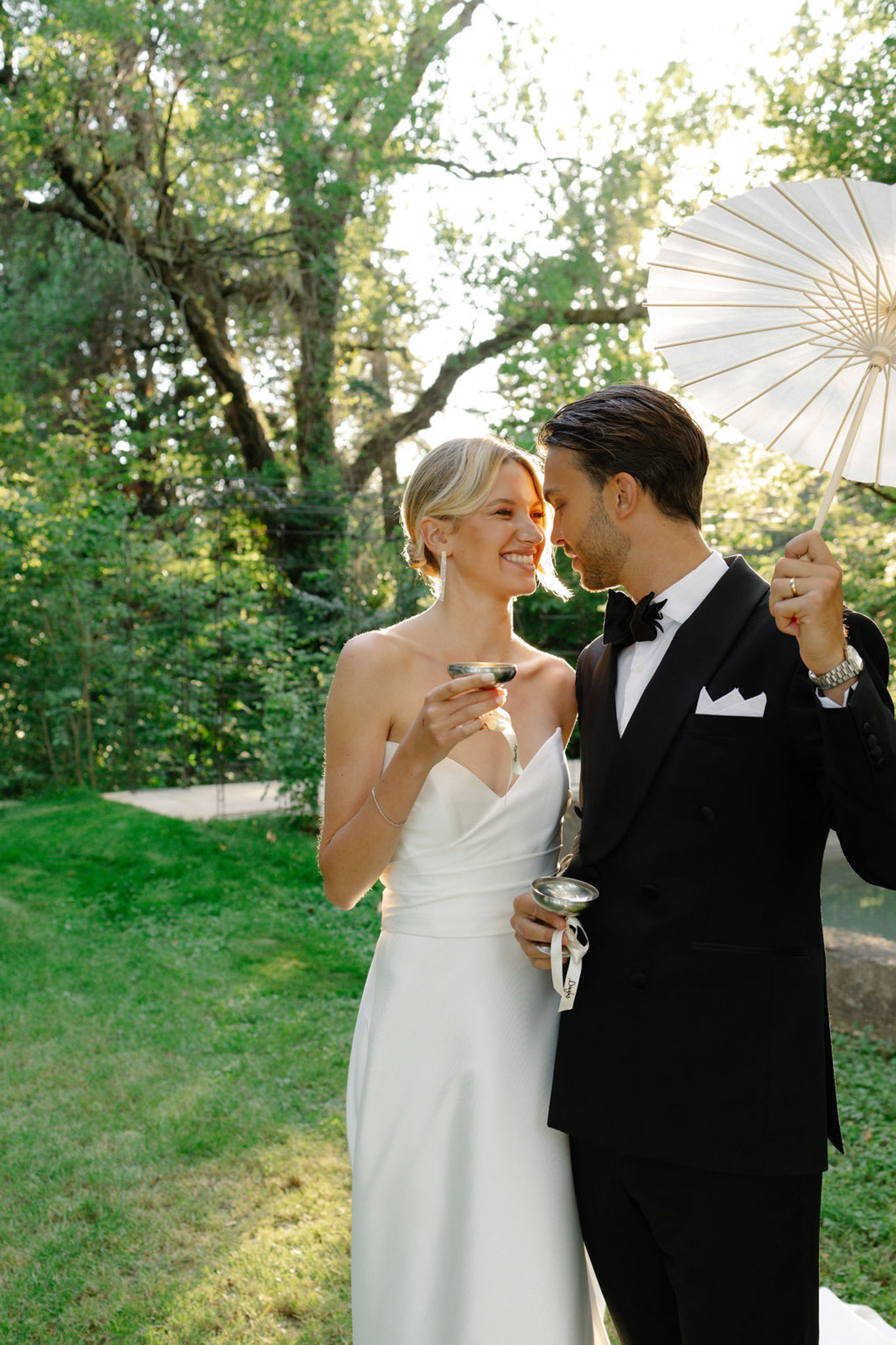Bride and groom sharing a candid moment under trees during cocktail hour, groom holding cream parasol