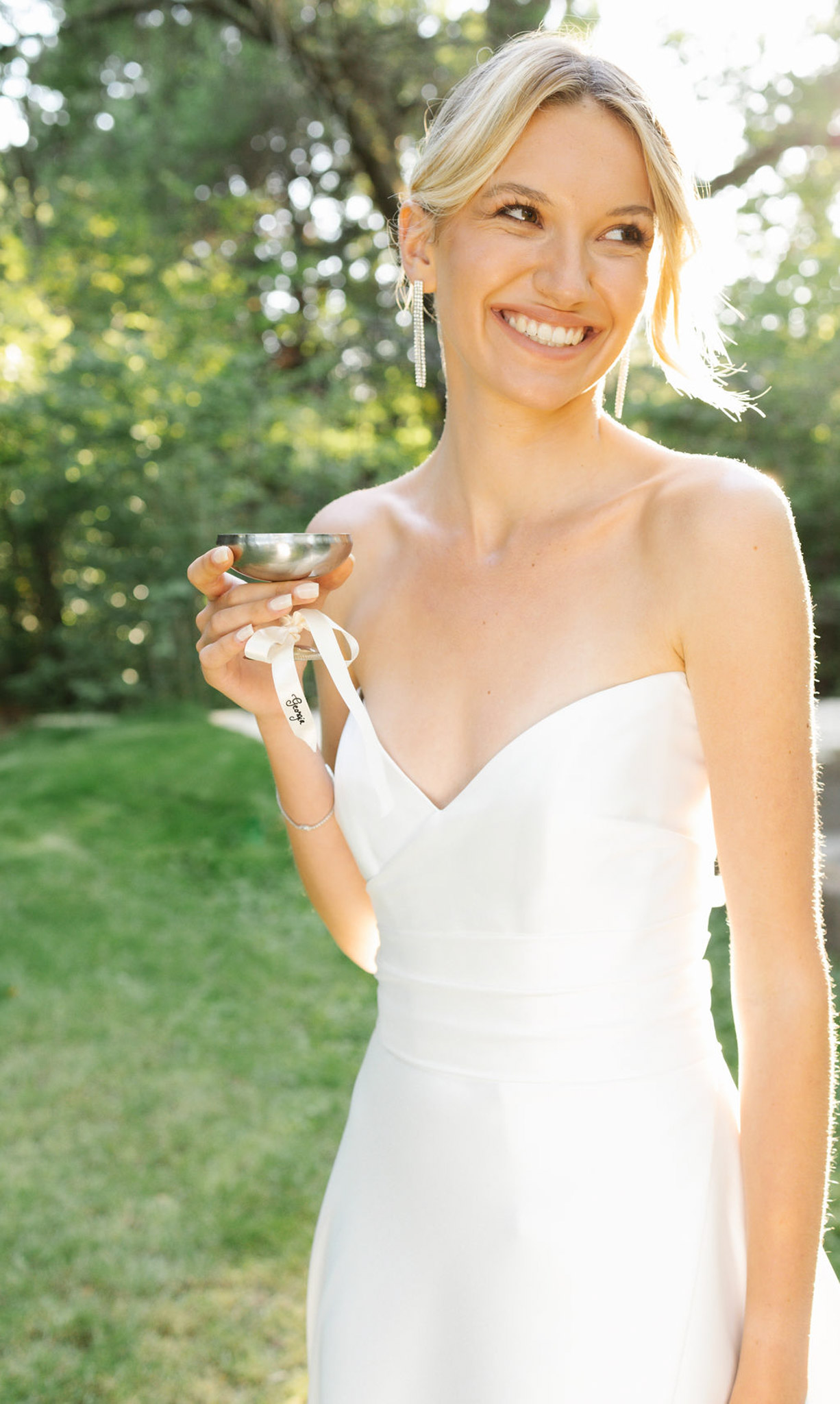 Bride in strapless ivory gown holding champagne coupe with ribbon bow in a sunlit garden setting