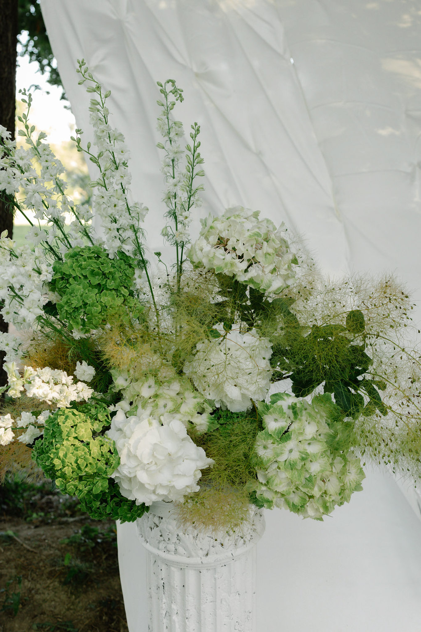 White hydrangeas, roses, baby's breath and astilbe floral arrangement attached to sheer white fabric drape on stone pedestal