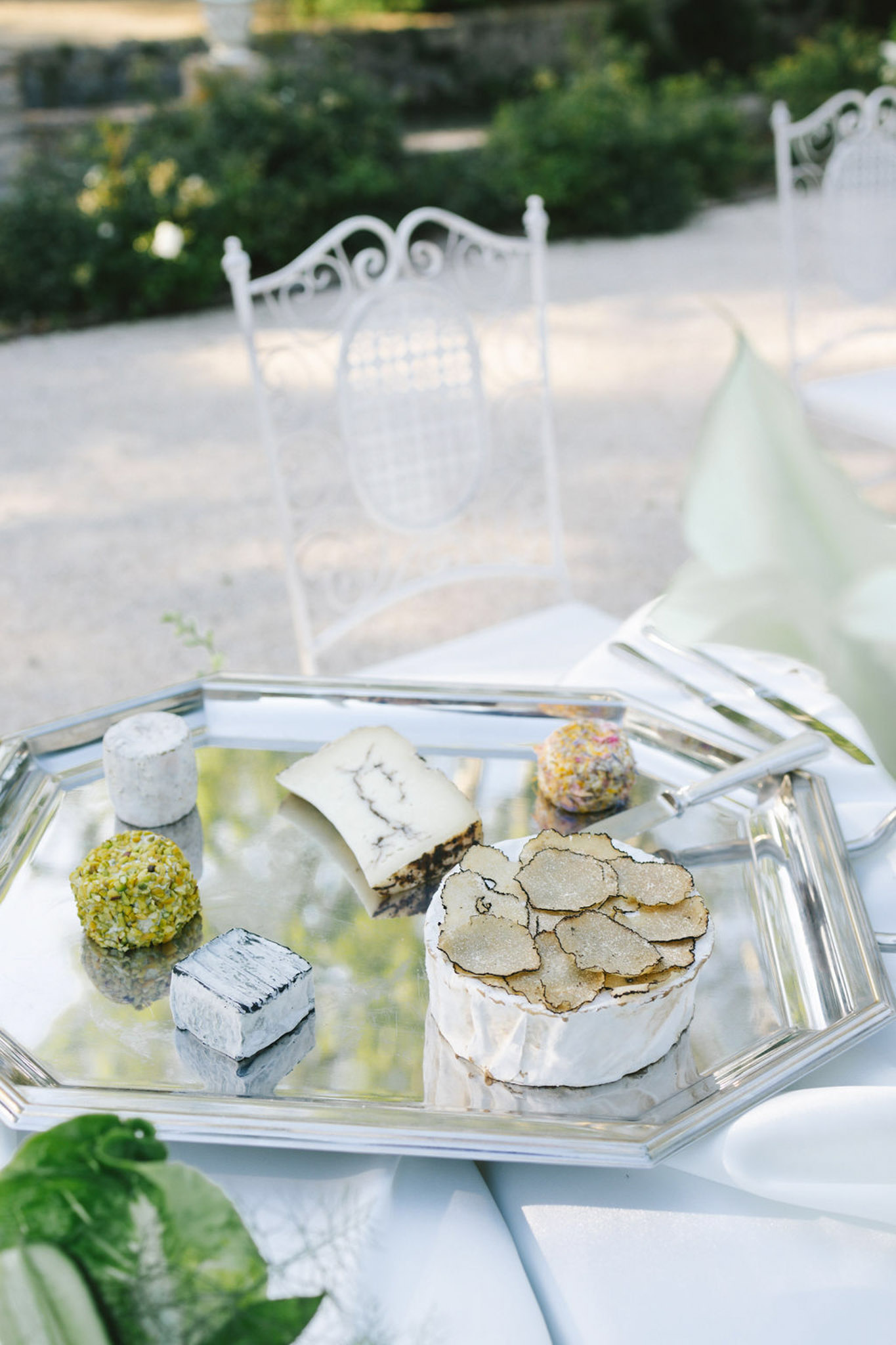 Gourmet cheese board on mirrored silver tray with five varieties, fresh herbs, and serving utensils at outdoor reception