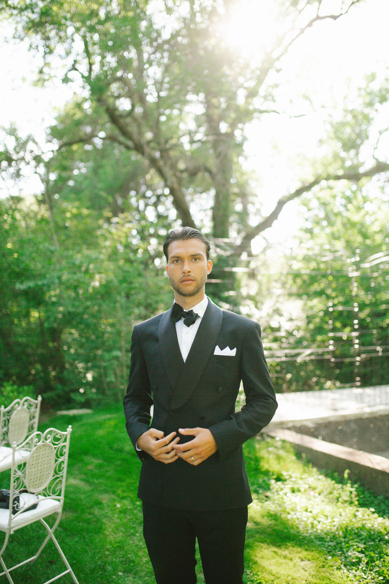 Groom in black double-breasted tuxedo with white pocket square stands on manicured lawn with ornate white garden chairs