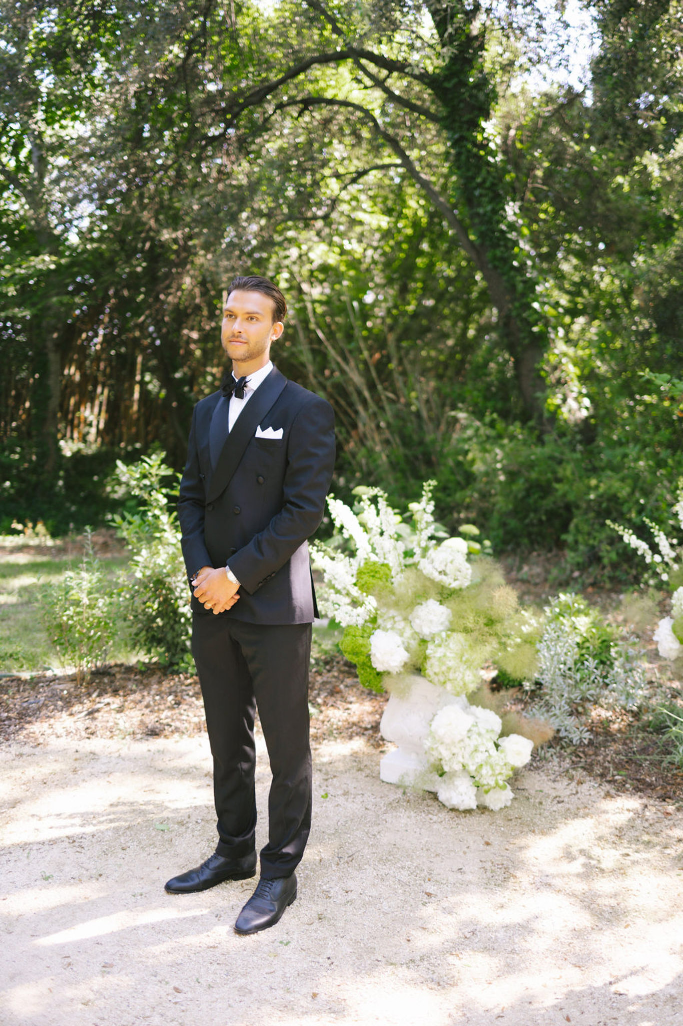 Groom in navy double-breasted suit and bow tie standing on a tree-lined garden path with hydrangea arrangements