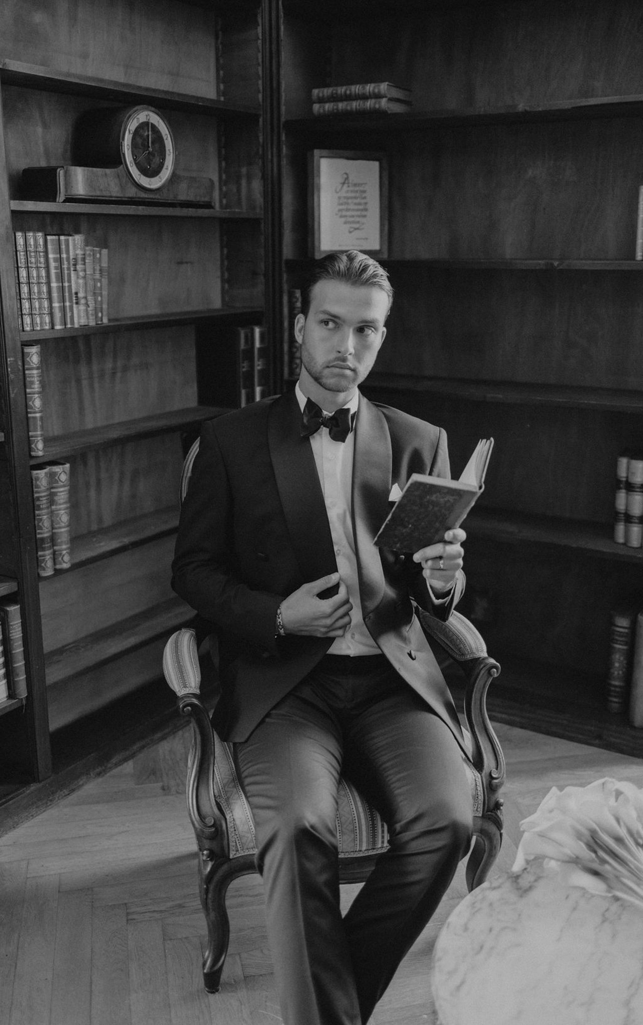 Black and white groom portrait seated in library holding book in tuxedo with bow tie