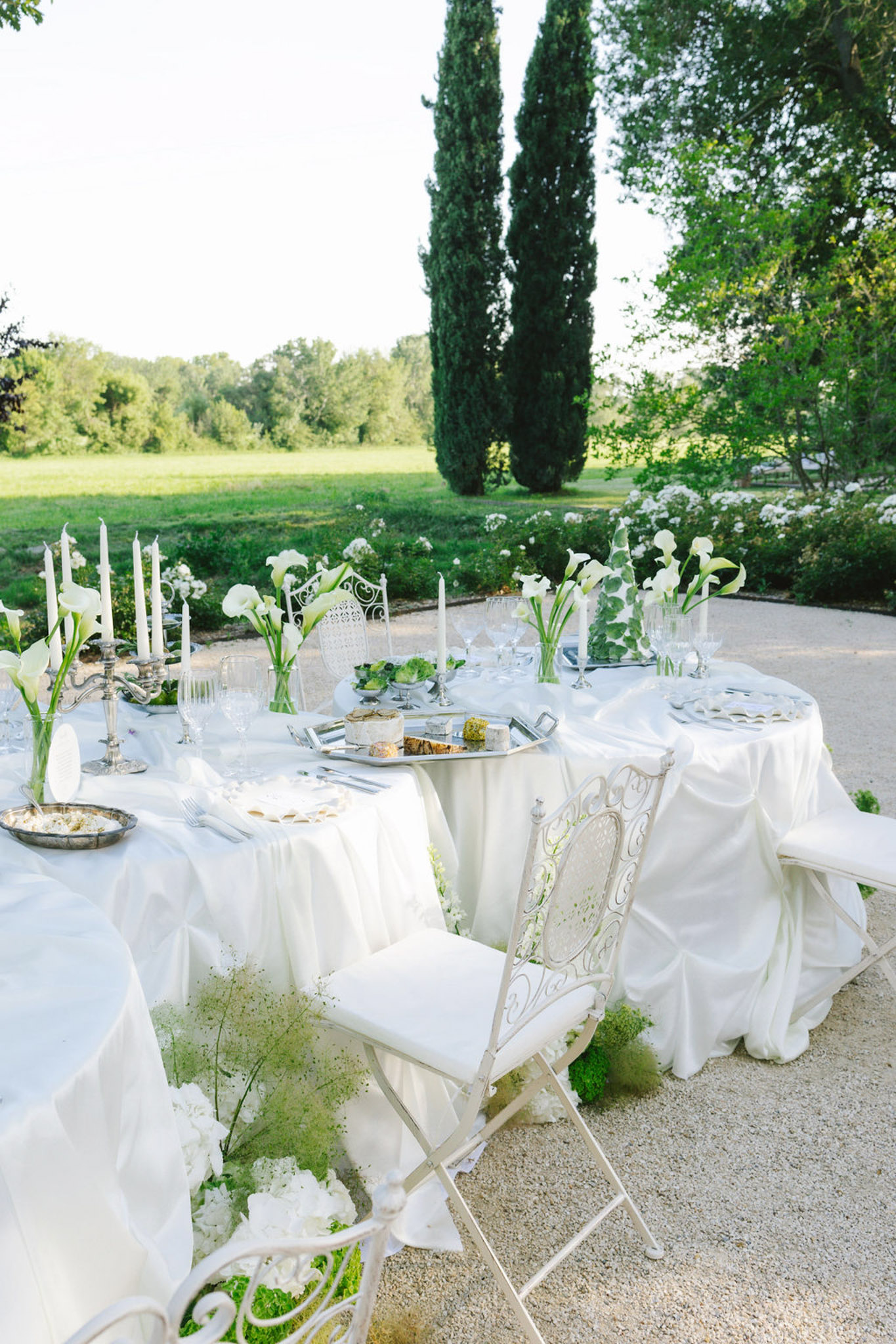 Outdoor reception tables on stone terrace with ivory linens, white calla lilies, carved white chairs, cypress trees