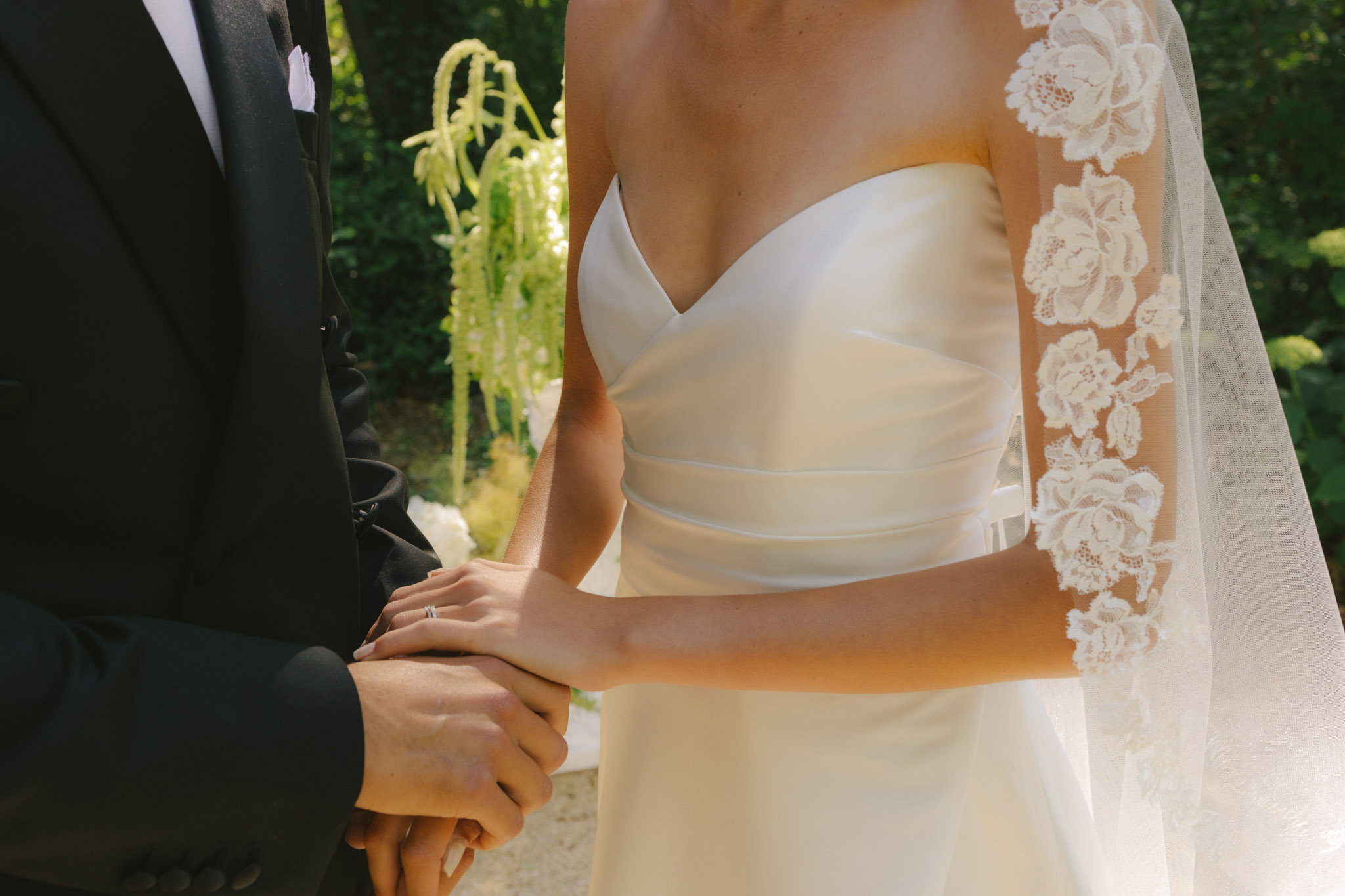 Close-up of bride and groom holding hands during ceremony, bride in lace appliquÃ© veil and ruched ivory gown