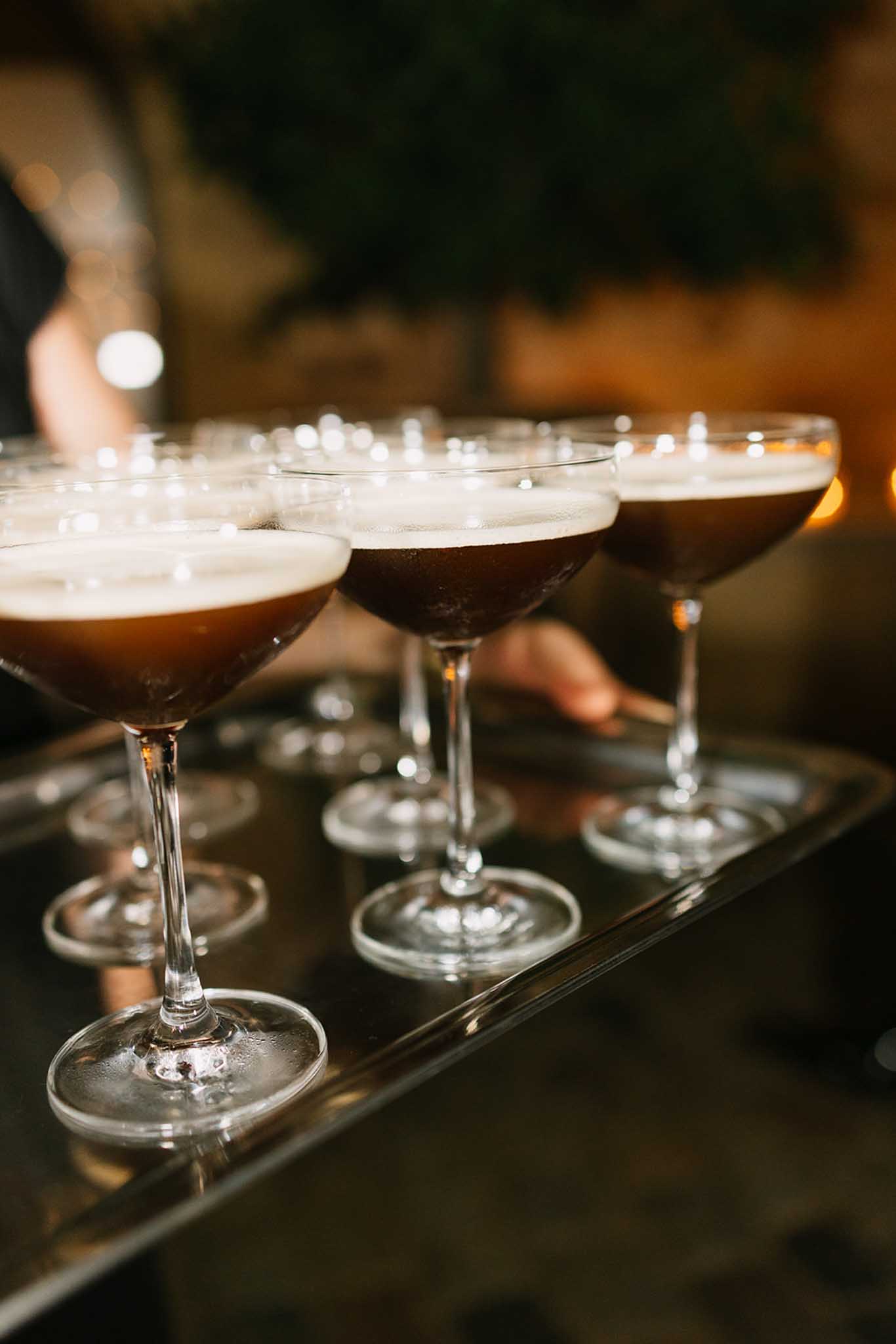 Row of coupe glasses filled with dark brown cocktails topped with cream foam on a black tray at a cocktail hour service