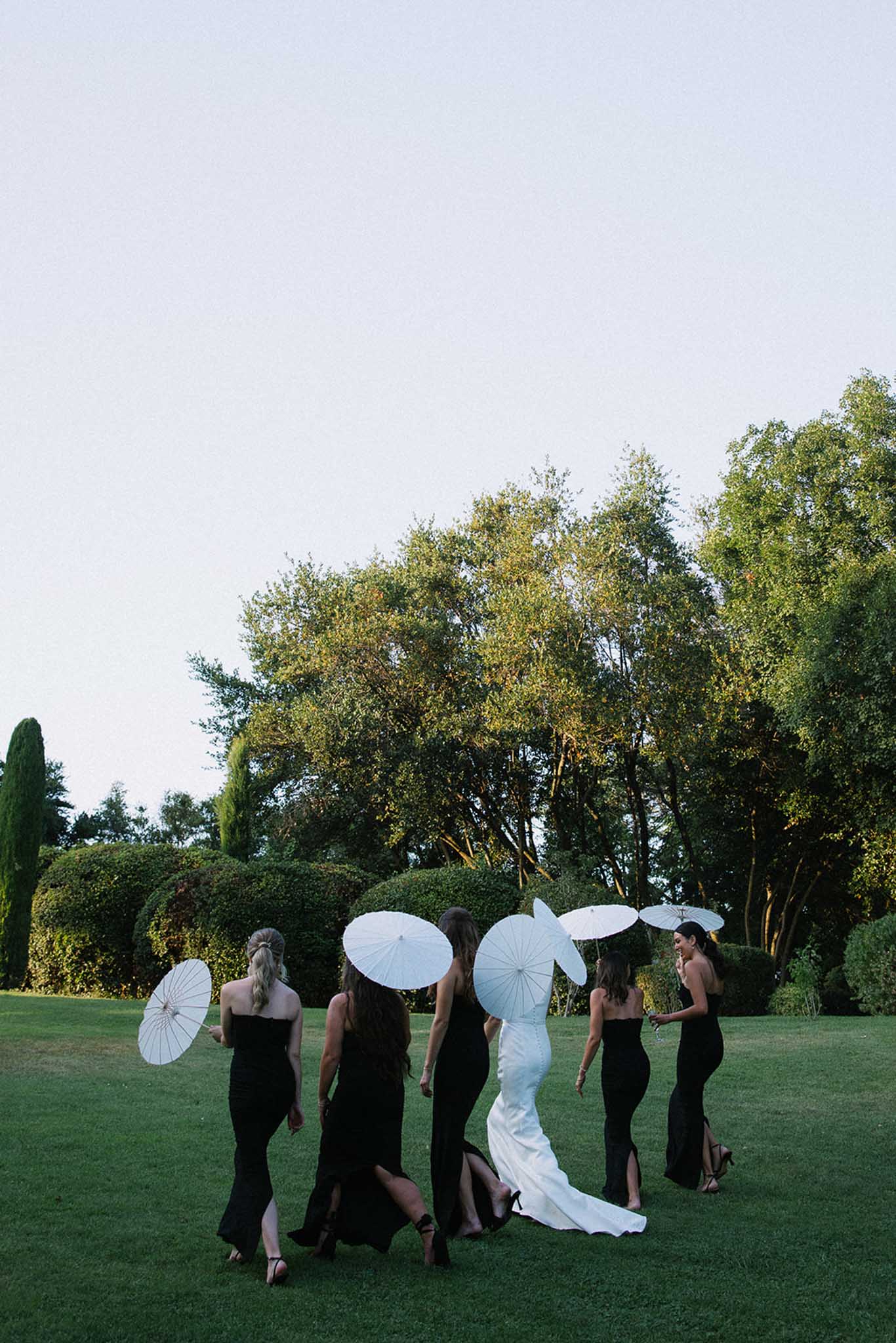 Bride and five bridesmaids in black dresses walking across a manicured lawn holding white parasols