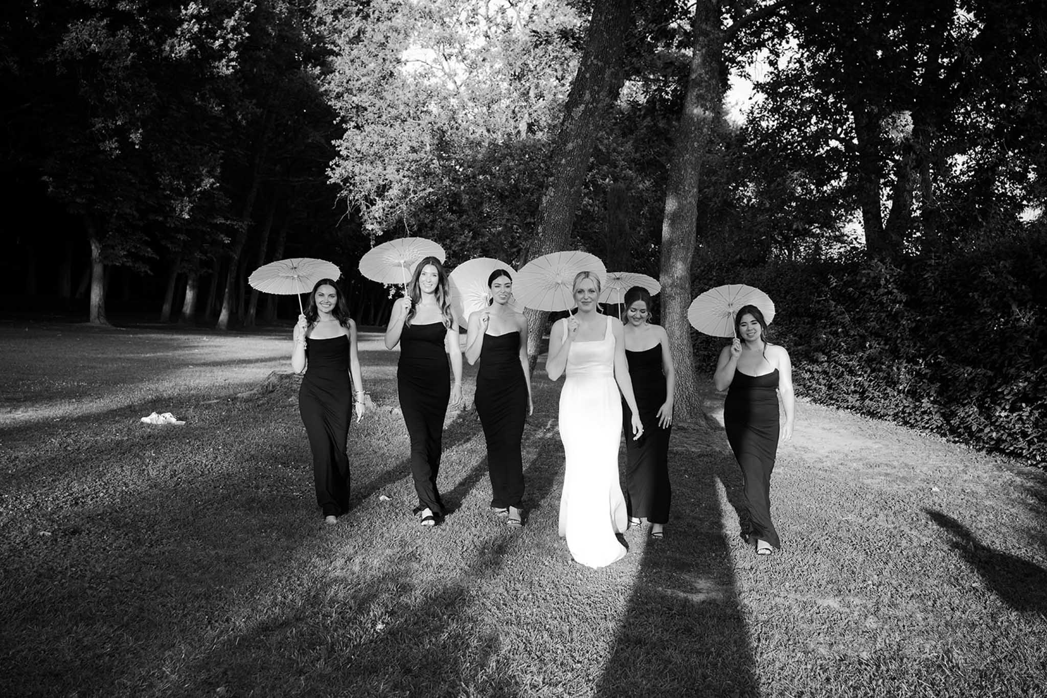 Bride in white and five bridesmaids in black walk toward camera holding white umbrellas on tree-lined path
