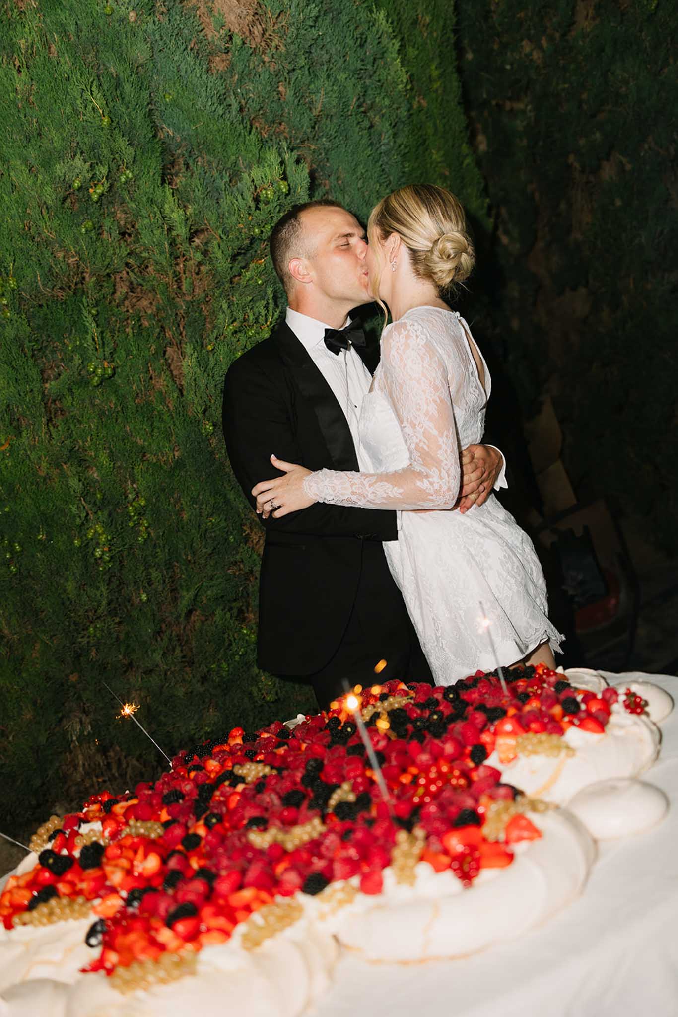 Bride and groom kiss during cake cutting with sparklers at nighttime reception in front of moss wall backdrop
