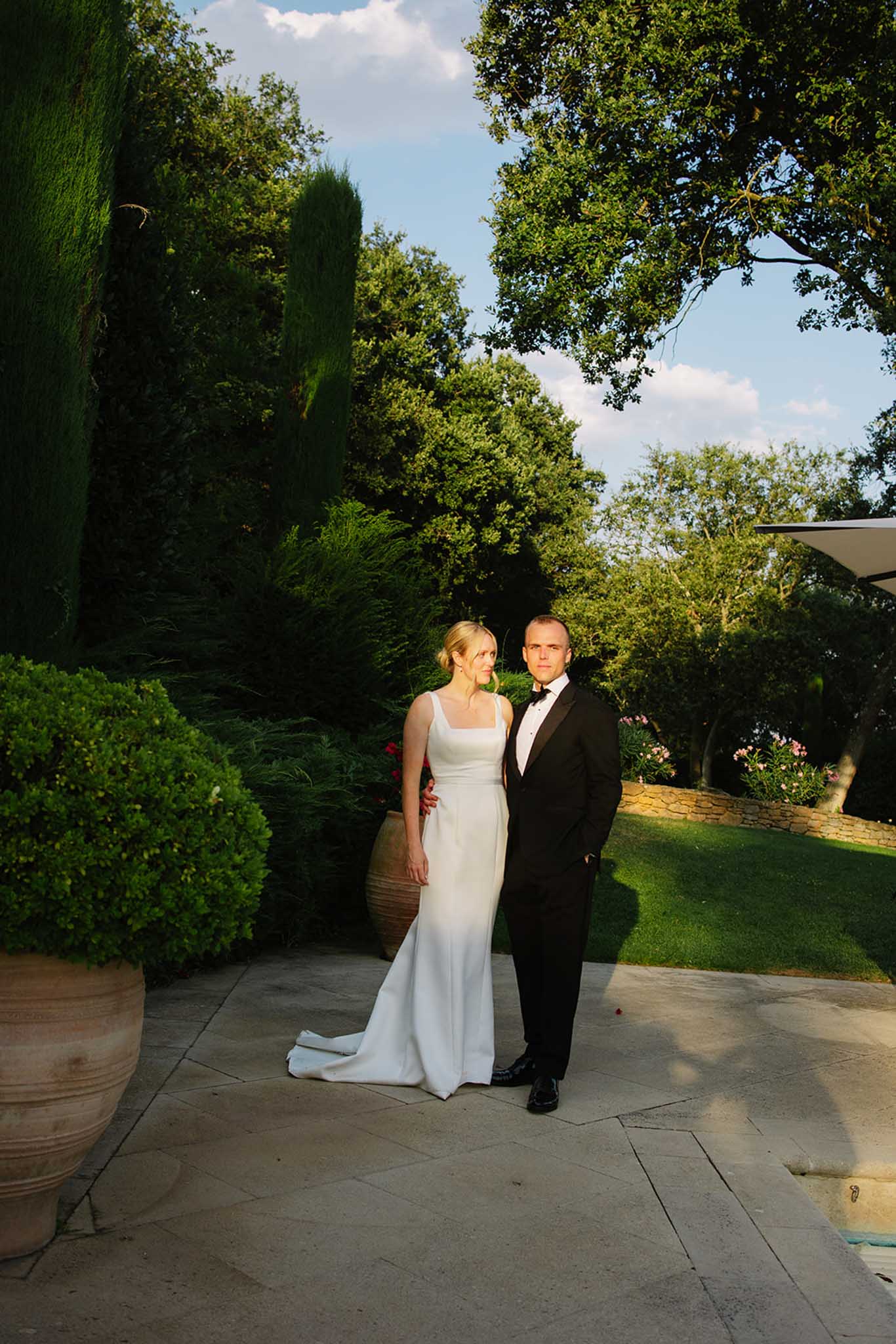 Bride in ivory column gown and groom in black tuxedo standing on stone patio, framed by cypress trees and boxwood hedges