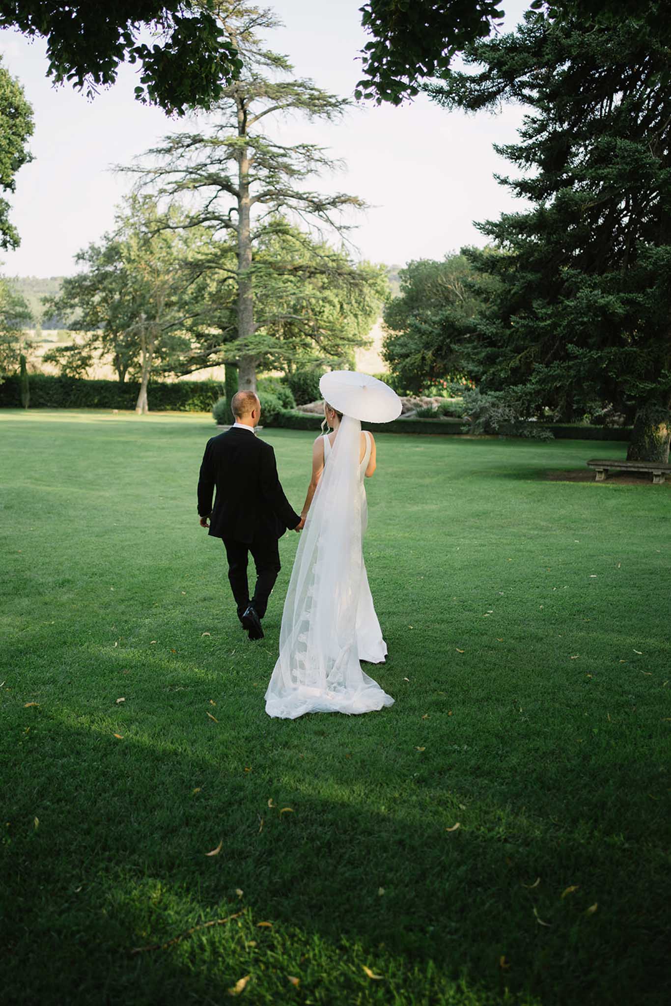 Bride and groom walking hand-in-hand across manicured lawn photographed from behind