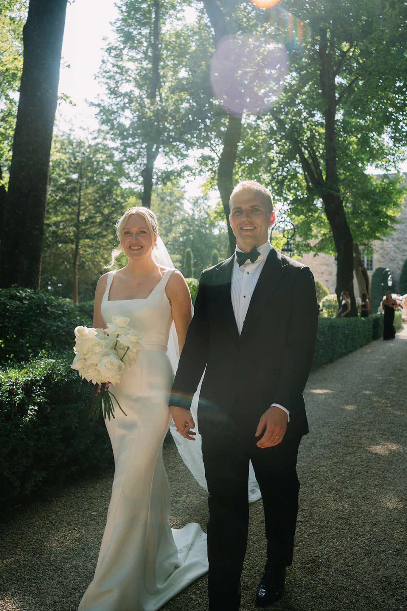 Bride and groom walking arm-in-arm down a tree-lined garden pathway with dappled sunlight