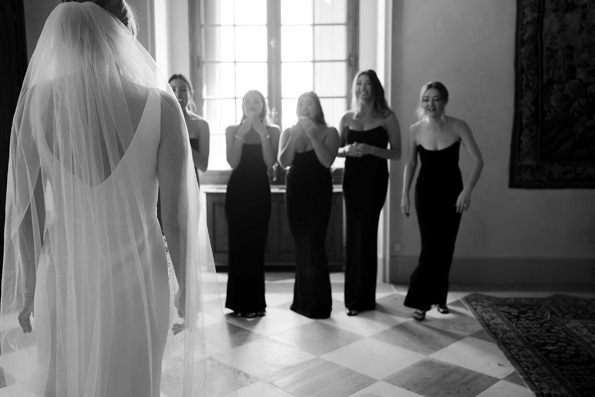Bride seen from behind in wedding dress and tulle veil faces five bridesmaids in black strapless gowns