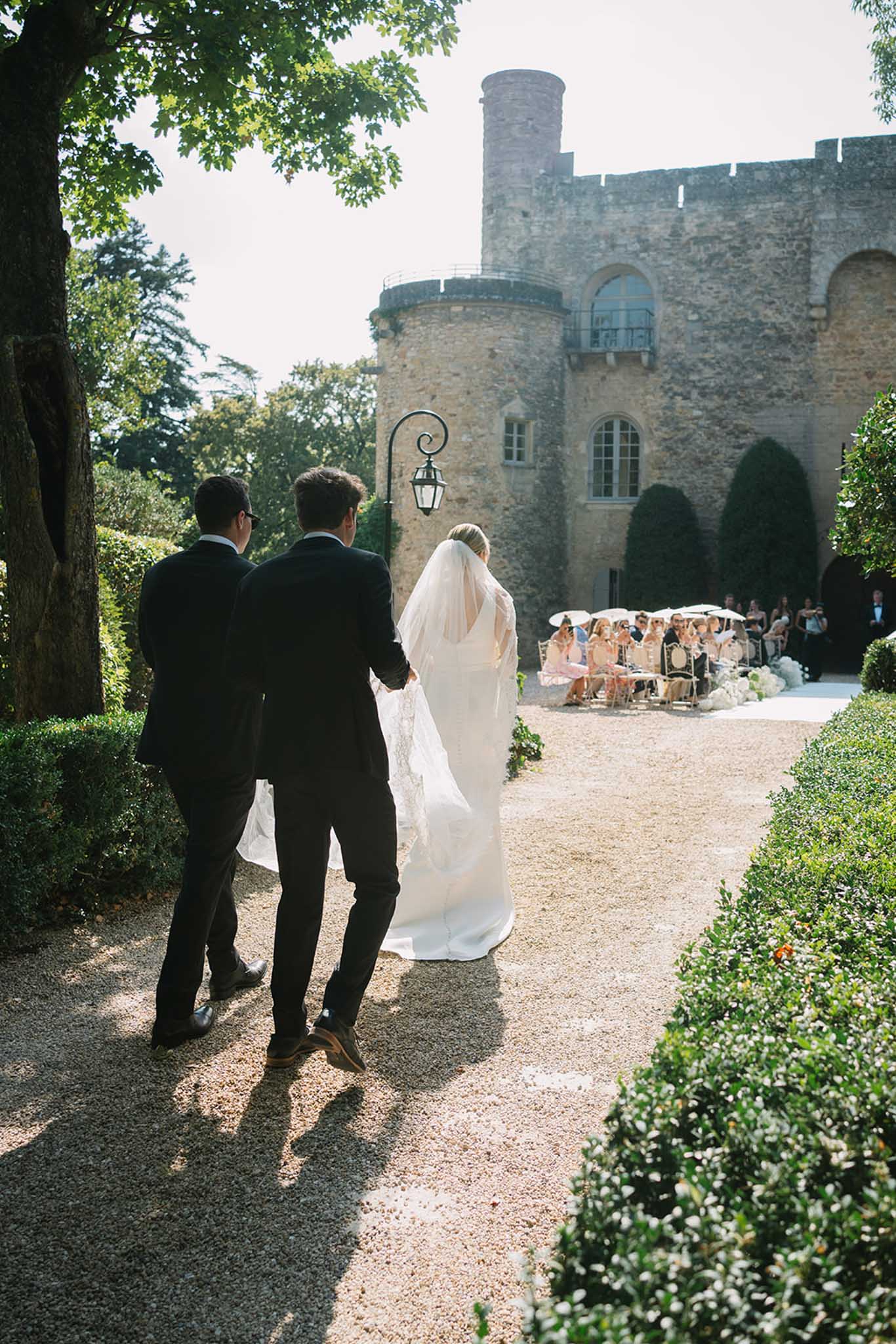 Bride and groom walking toward ceremony setup in front of stone castle with crenellated towers and seated guests